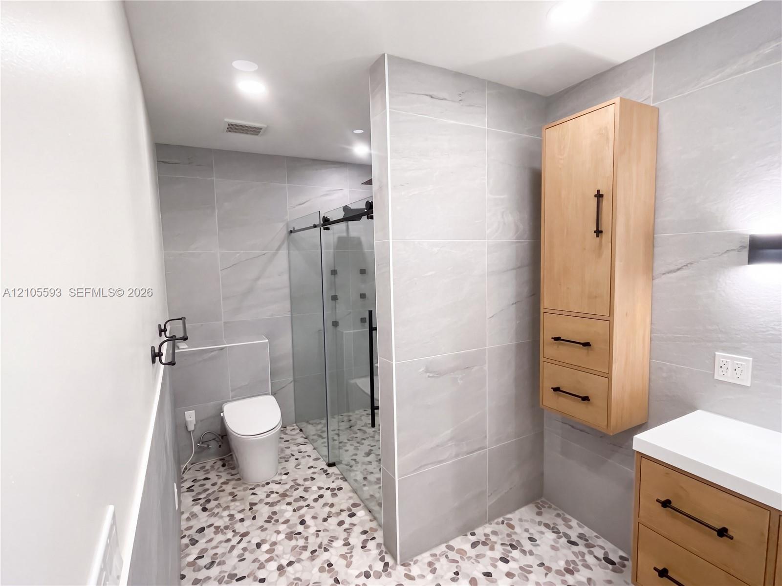 Bathroom featuring large-format gray wall tile, a glass-enclosed shower with multiple spray heads, pebble-style flooring, wall-mounted wood-finish cabinetry, and a white countertop