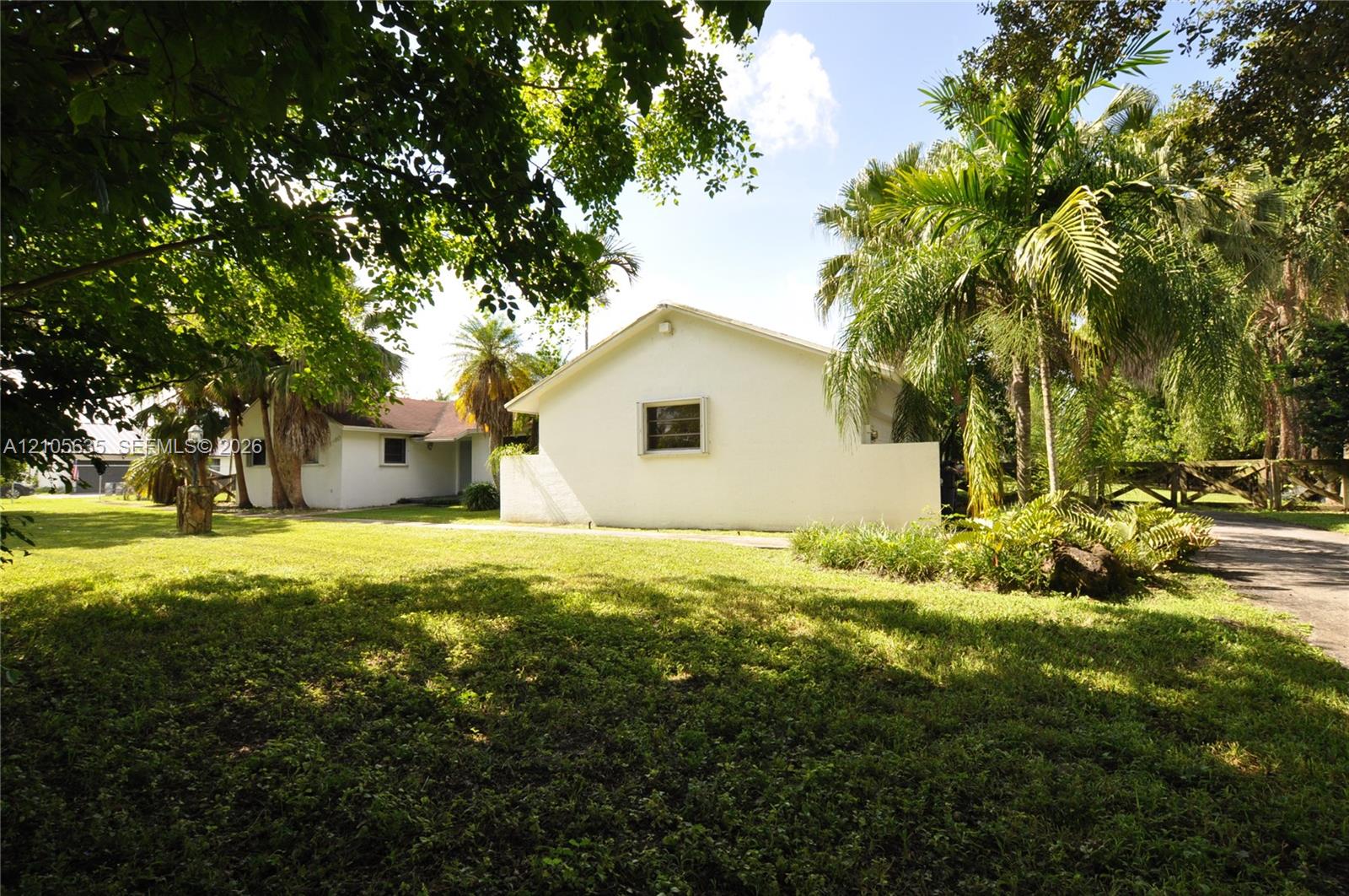 Stucco exterior residence featuring a sprawling lawn, mature palm trees, perimeter wooden fencing, and a paved driveway