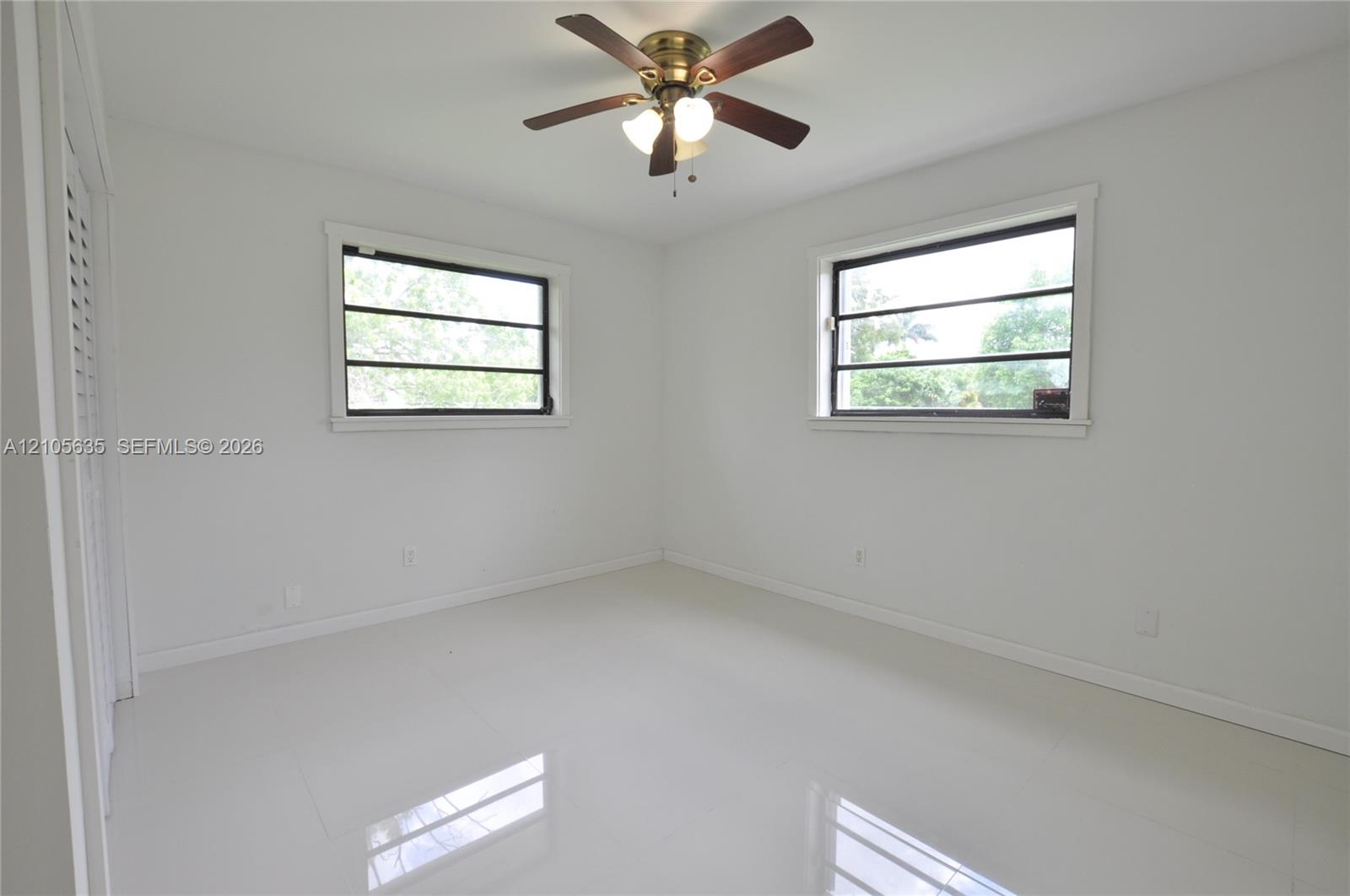 Neutral interior featuring large-format tile flooring, dual windows, a ceiling fan with integrated lighting, and louvered closet doors