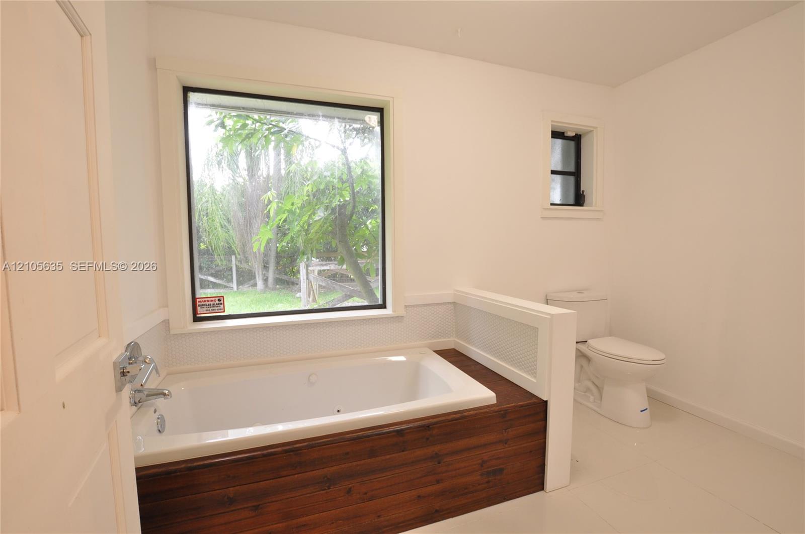 Built-in soaking tub with wood-finish surround, large window with exterior landscape, tile flooring, and crisp white wall finish