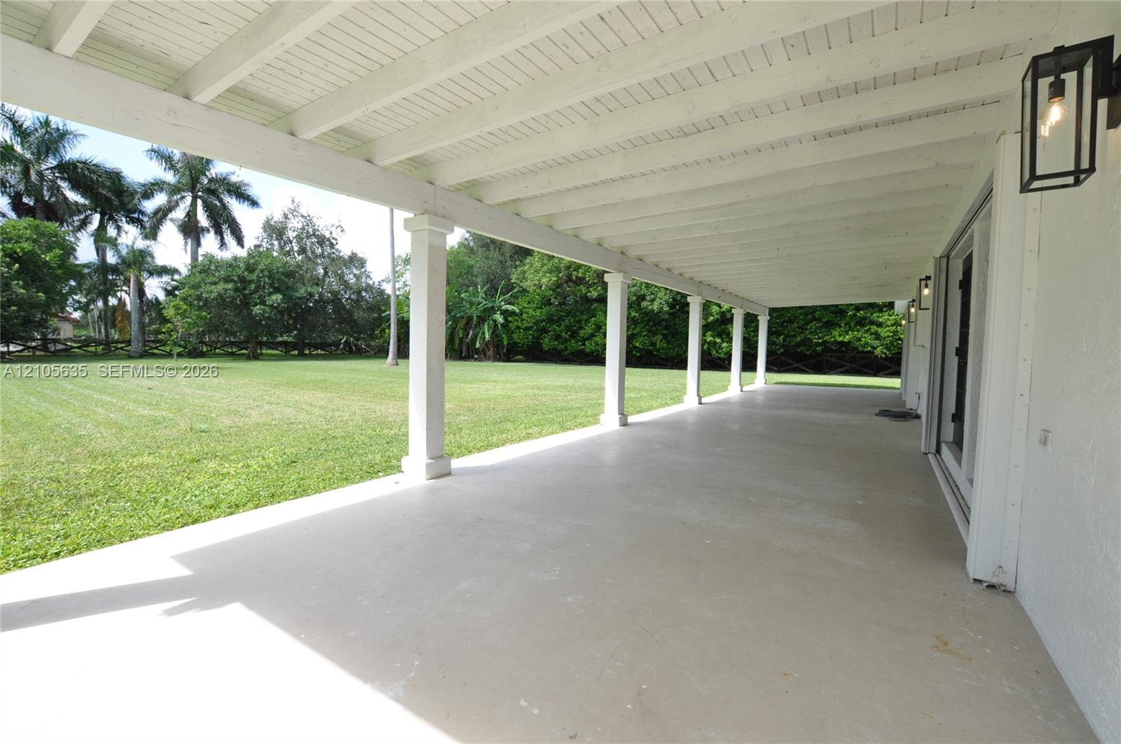 Covered patio featuring a painted wood-beam ceiling, structural columns, sleek concrete flooring, and minimalist wall-mounted light fixtures, overlooking an expansive manicured lawn