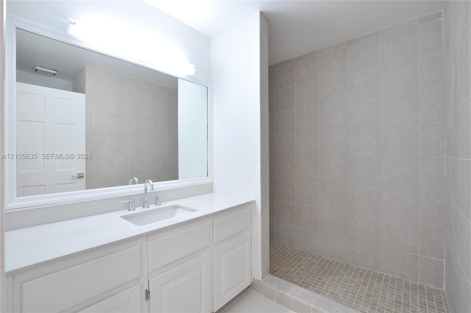 Bathroom featuring a white vanity cabinet with a solid surface countertop, undermount sink, expansive wall-mounted mirror, and a walk-in shower with floor-to-ceiling tile surround