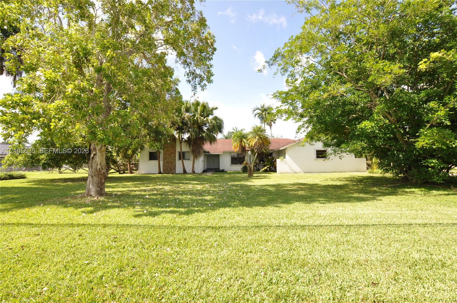Expansive lawn with mature trees and palms, featuring a white exterior residence with stone-clad masonry detail and a shingled roof