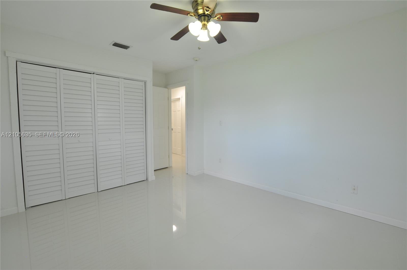 Neutral-toned tile flooring, white walls, and a multi-panel closet with white louvered doors, accented by a central ceiling fan with integrated lighting