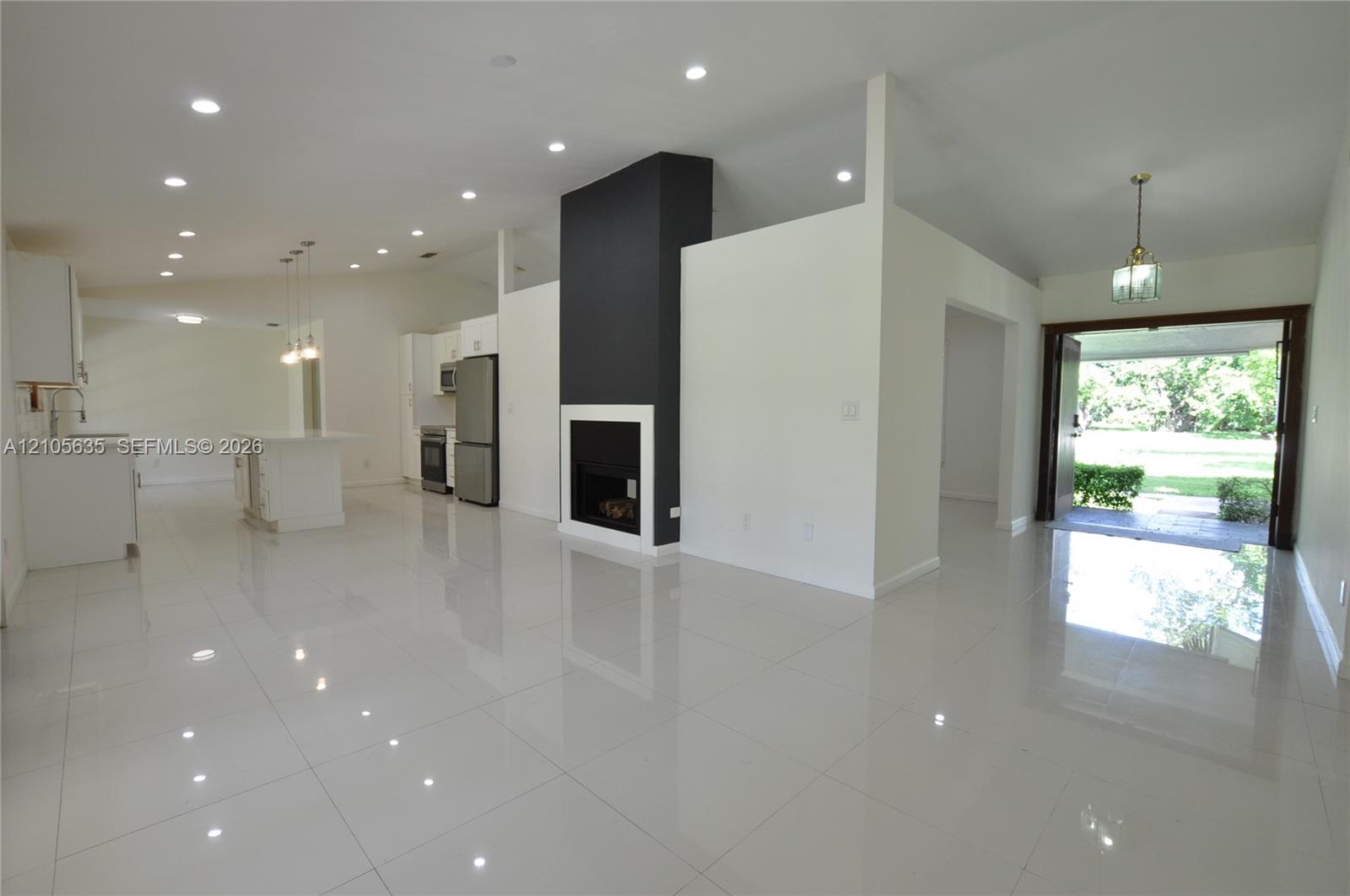 Open-concept interior featuring large-format polished tile flooring, vaulted ceilings with recessed lighting, a modern floor-to-ceiling fireplace with high-contrast surround, and stainless steel kitchen appliances