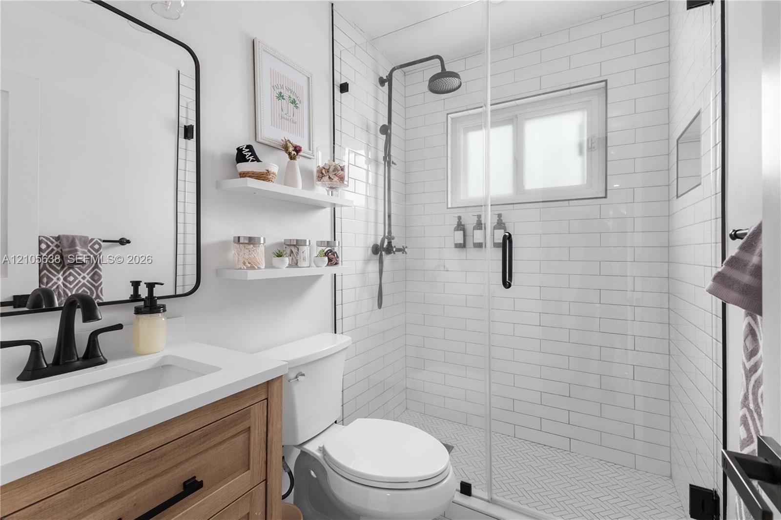 Bathroom featuring a wood-finish vanity with a white countertop, matte black fixtures, white subway tile shower enclosure with glass door, and wall-mounted shelving