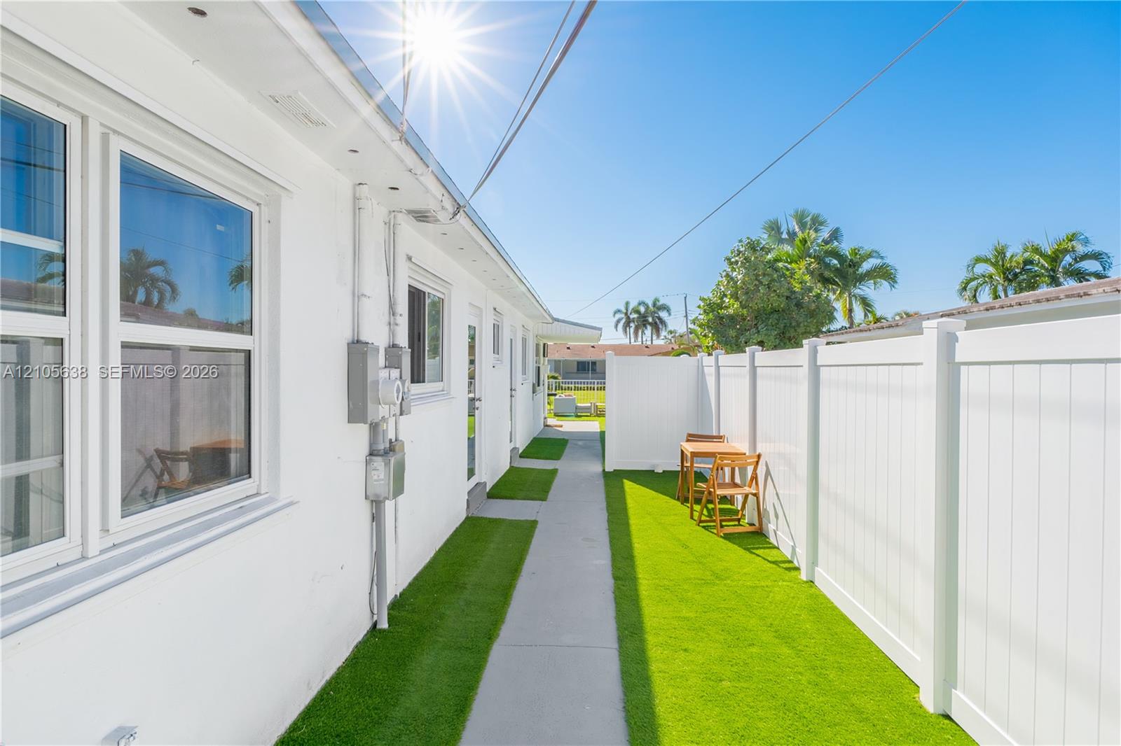 Exterior side yard featuring white stucco siding, a concrete pathway, artificial turf ground cover, and a full-height white vinyl privacy fence