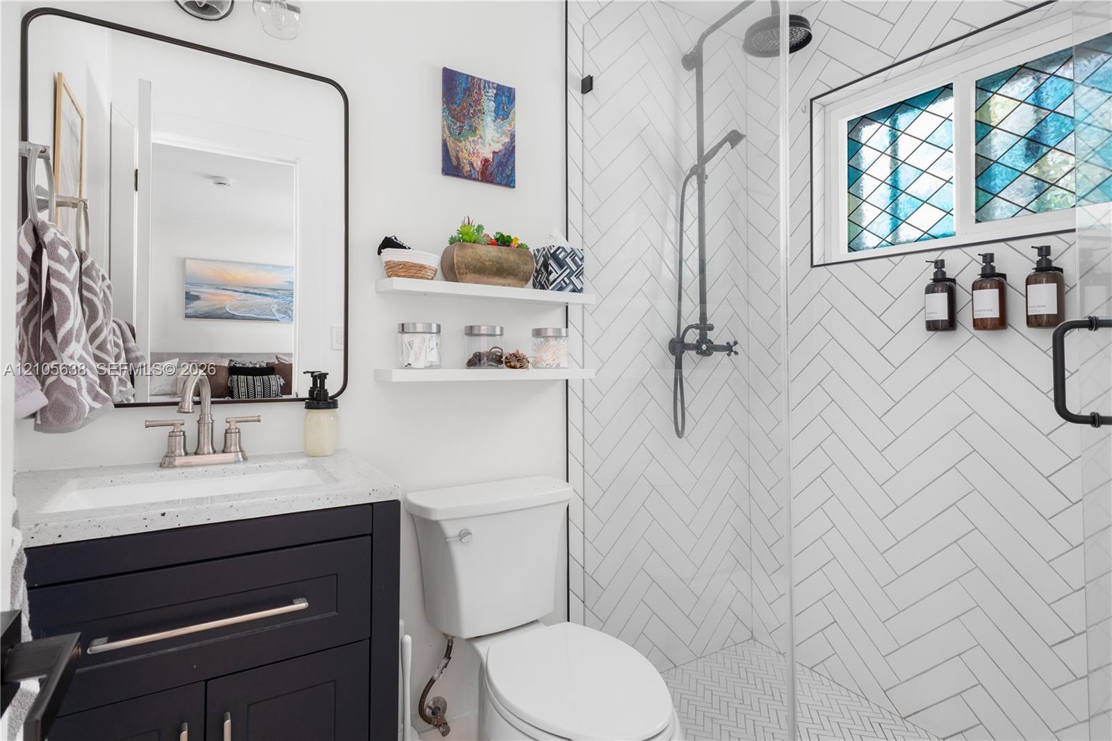Bathroom featuring a dark-finish vanity with a speckled countertop, a walk-in shower with white herringbone-patterned wall tile, a glass shower enclosure, and a window with decorative leaded-glass detailing