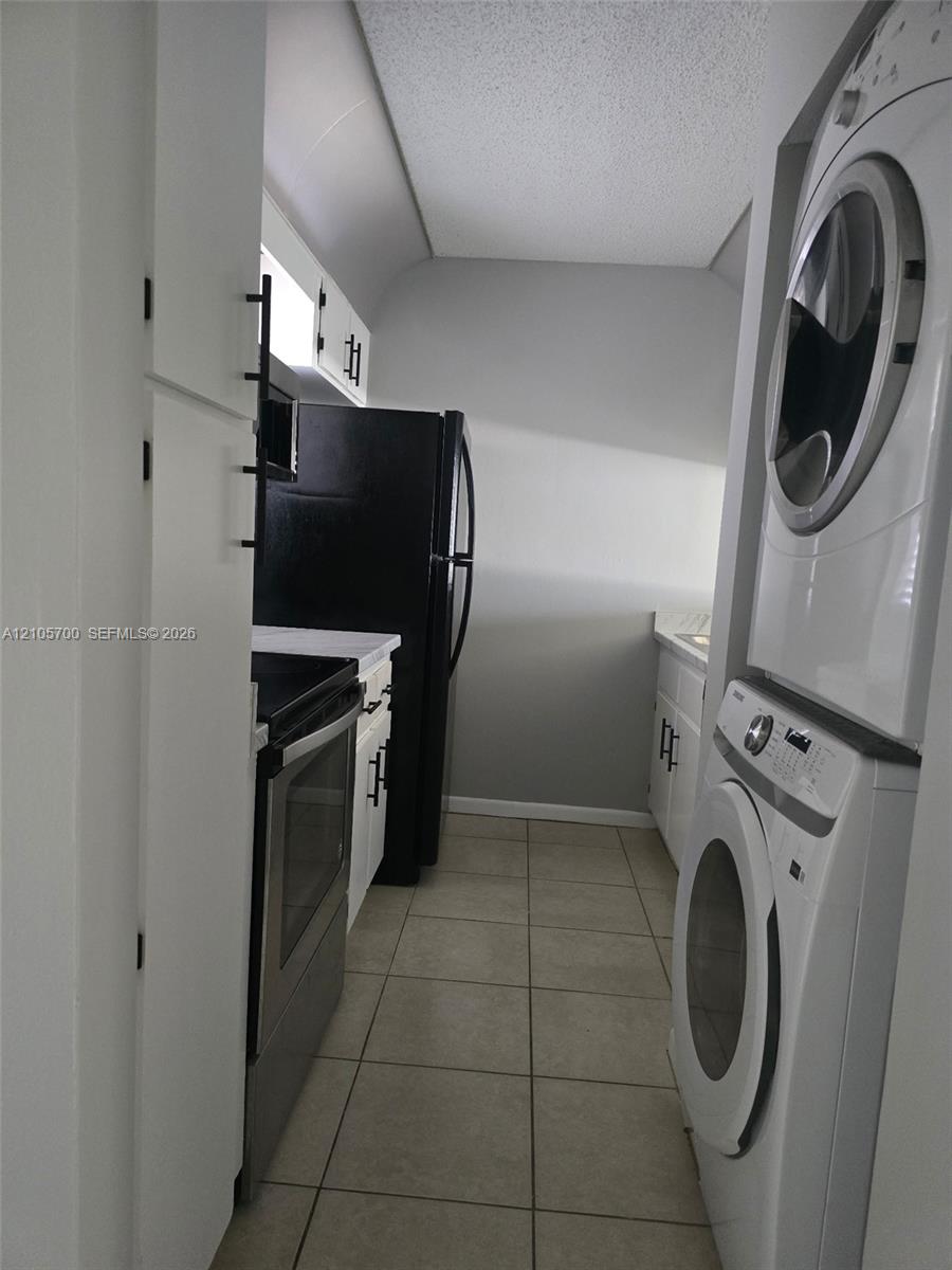 Utility space featuring large-format tile flooring, stacked front-load laundry appliances, white cabinetry with matte black hardware, and an integrated refrigerator and range