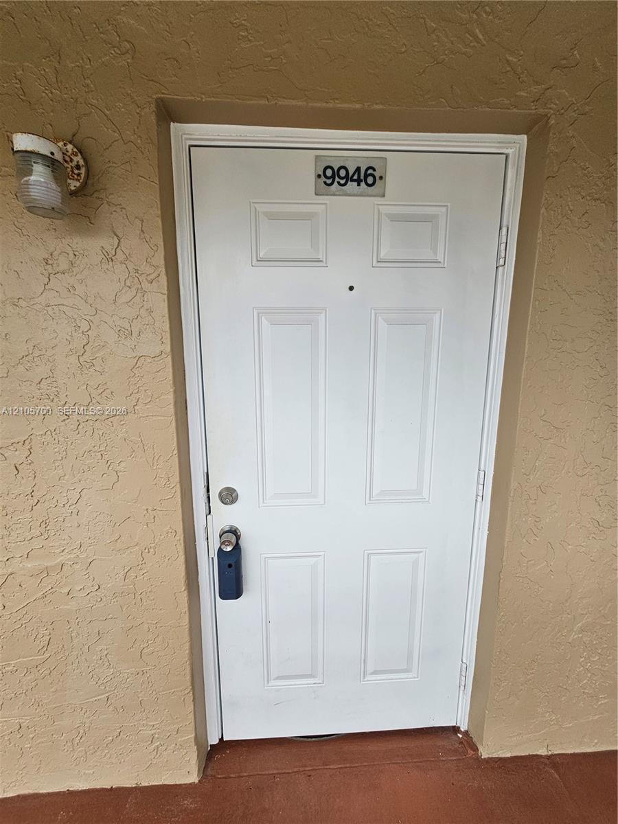 White six-panel entry door featuring a center-mounted peephole, metal address plaque, and wall-mounted exterior light fixture