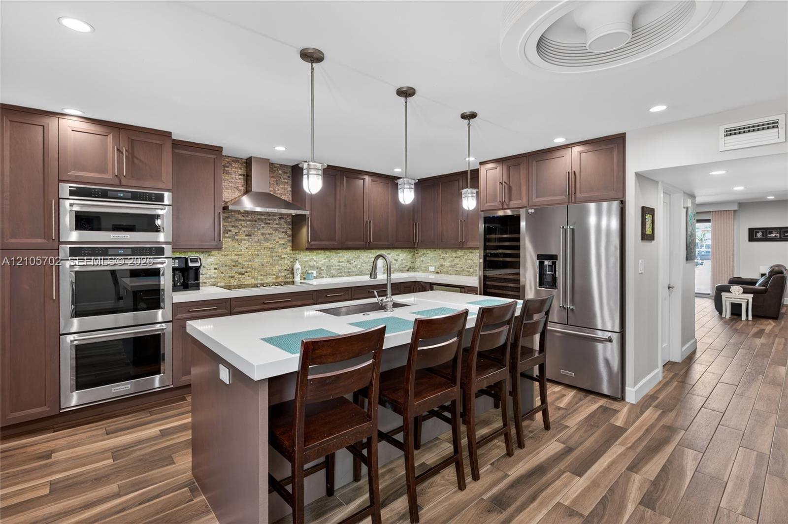 Kitchen featuring wood-finish flooring, stone tile backsplash, stainless steel built-in double ovens, quartz island countertop, and glass-front wine refrigerator