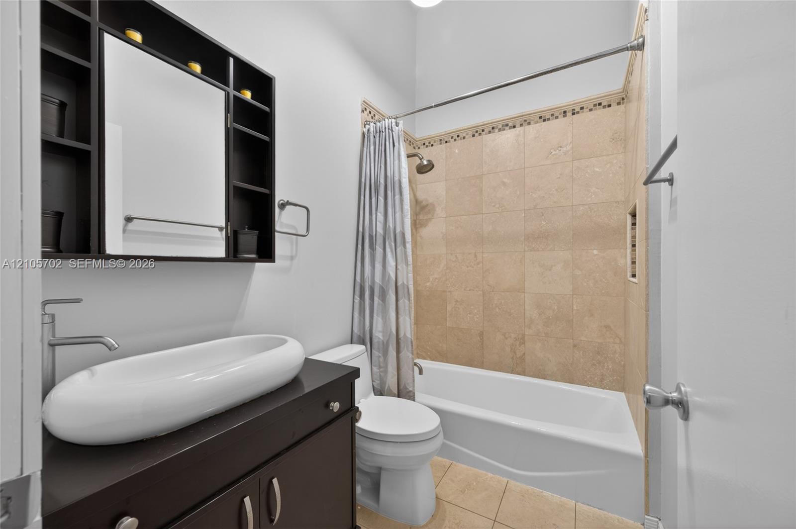 Dark wood-finish vanity with an oval vessel sink, wall-mounted medicine cabinet with integrated shelving, tiled tub surround with decorative inlay, and high-ceiling architecture