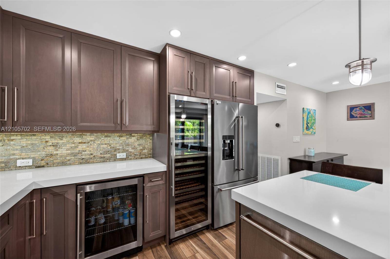 Kitchen featuring custom dark wood cabinetry, light-toned stone countertops, a multi-textured stone backsplash, stainless steel appliances including a built-in wine refrigerator, and wood-finish flooring