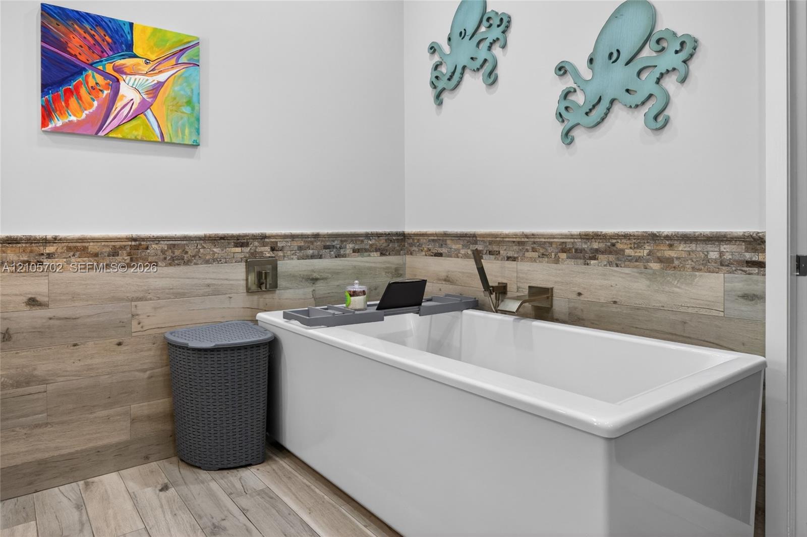 Deep soaking bathtub featuring a wall-mounted faucet, complemented by wood-finish wall paneling, stone-texture tile wainscoting, and wood-finish tile flooring