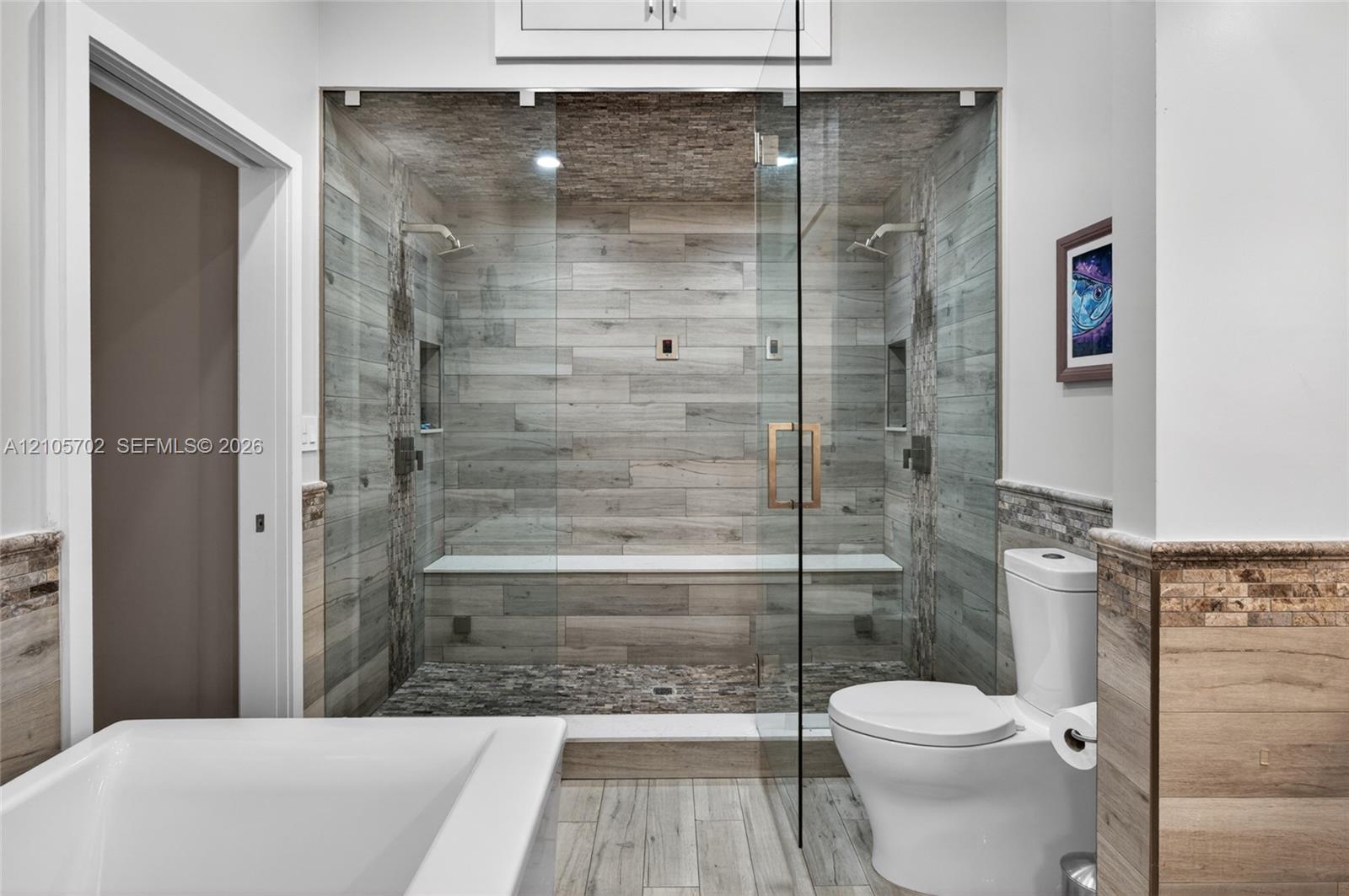 Walk-in shower featuring wood-finish wall tile, a built-in bench, dual overhead showerheads, recessed wall niches, and a frameless glass enclosure