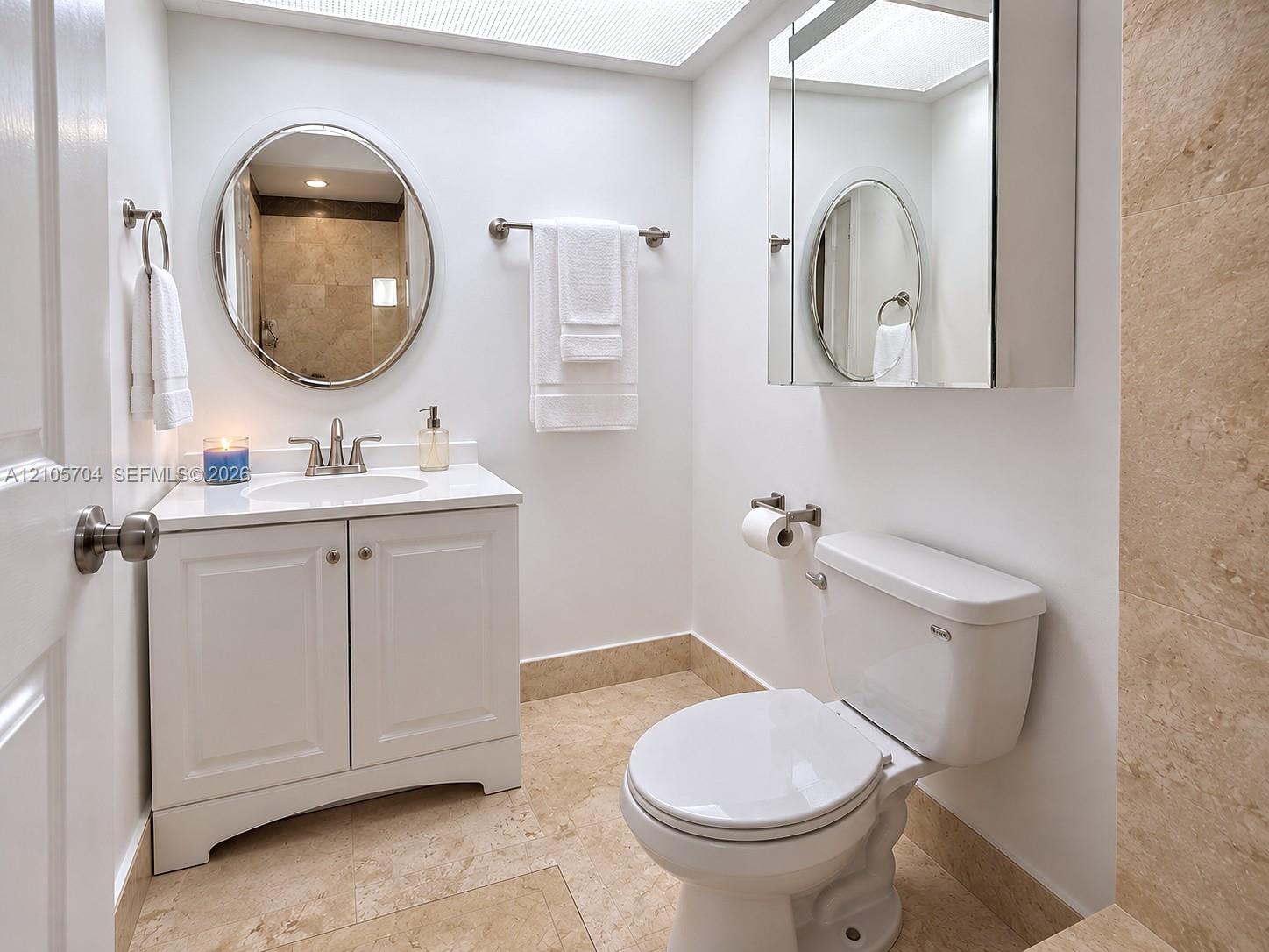 White vanity with a stone countertop, oval wall-mounted mirror, inset medicine cabinet, and light-toned stone tile flooring