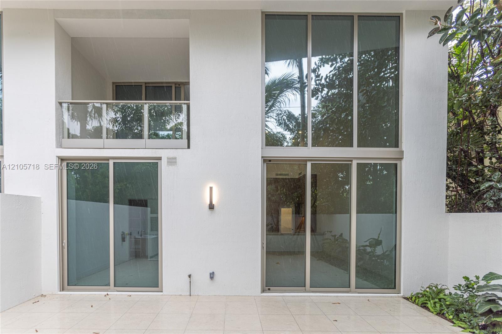 Exterior elevation featuring expansive two-story floor-to-ceiling windows, a covered balcony with a glass railing, large-format tile patio flooring, and wall-mounted minimalist lighting