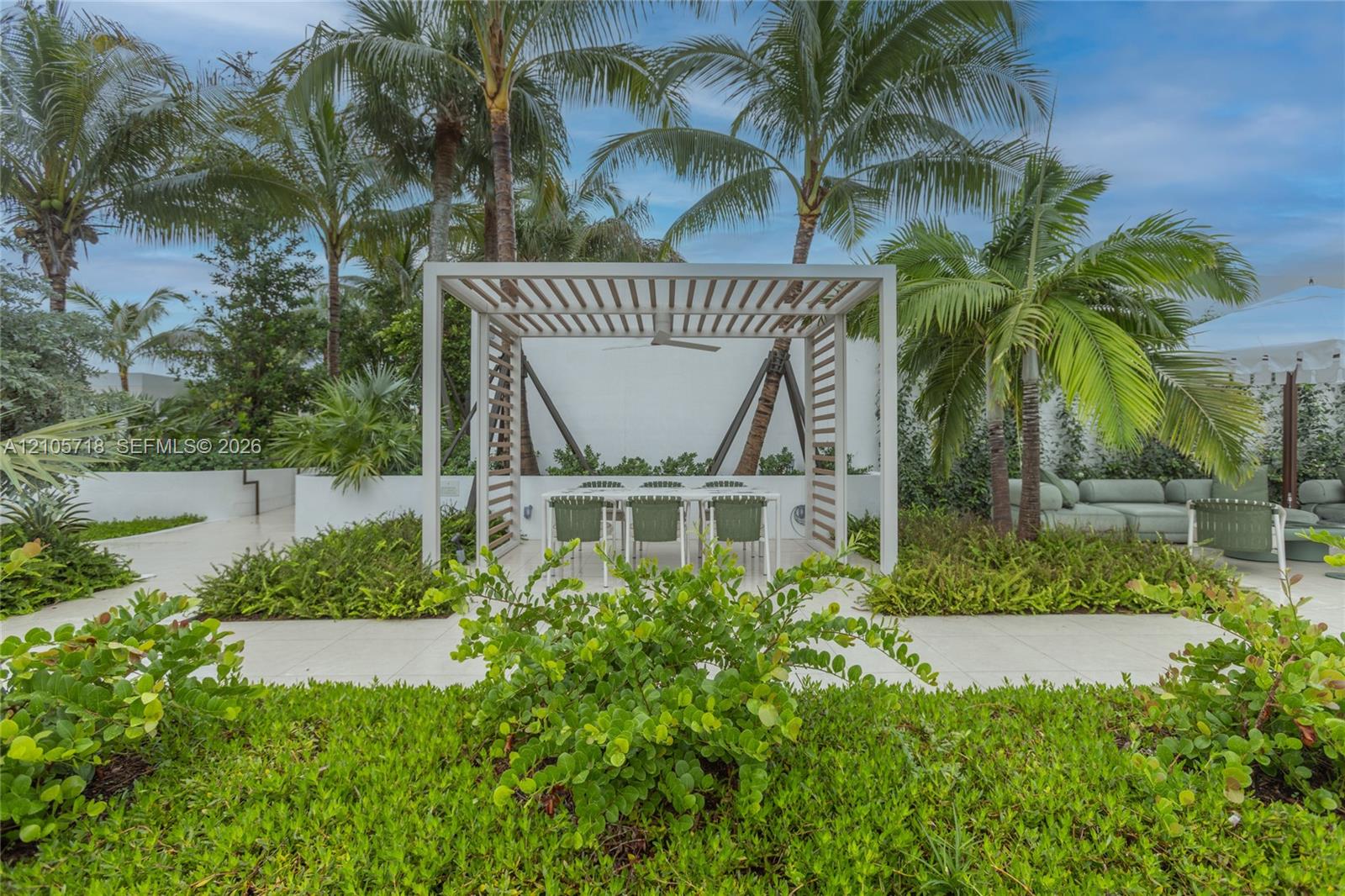 Outdoor patio featuring a white slatted pergola, integrated ceiling fan, light-colored paver flooring, raised garden beds, and mature palm trees