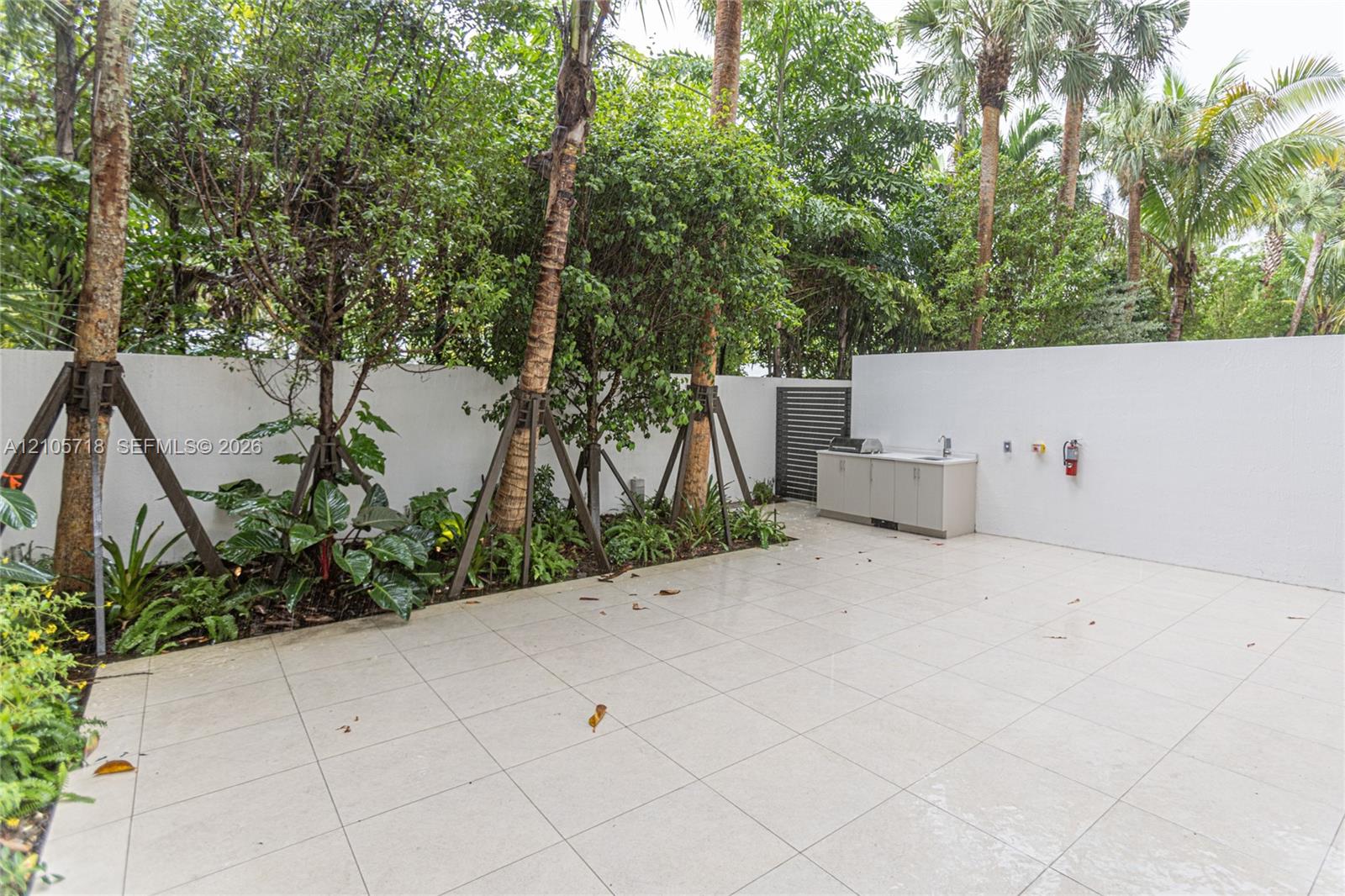 Private outdoor patio featuring light-toned tile flooring, built-in cabinetry with a sink, stainless steel grill, solid privacy walls, and mature perimeter landscaping
