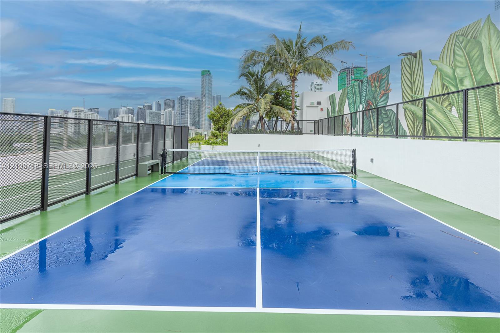 Rooftop pickleball court featuring a high-contrast blue and green acrylic surface, integrated netting, perimeter metal fencing, and decorative wall mural