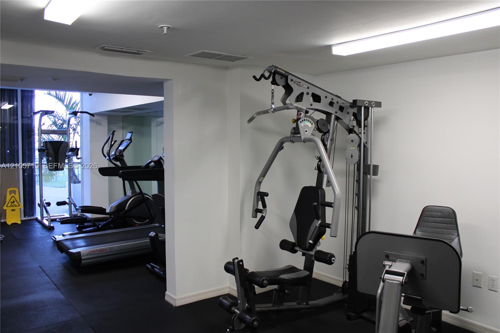 Fitness area featuring resilient rubber flooring, expansive wall-to-wall mirrors, and overhead fluorescent lighting