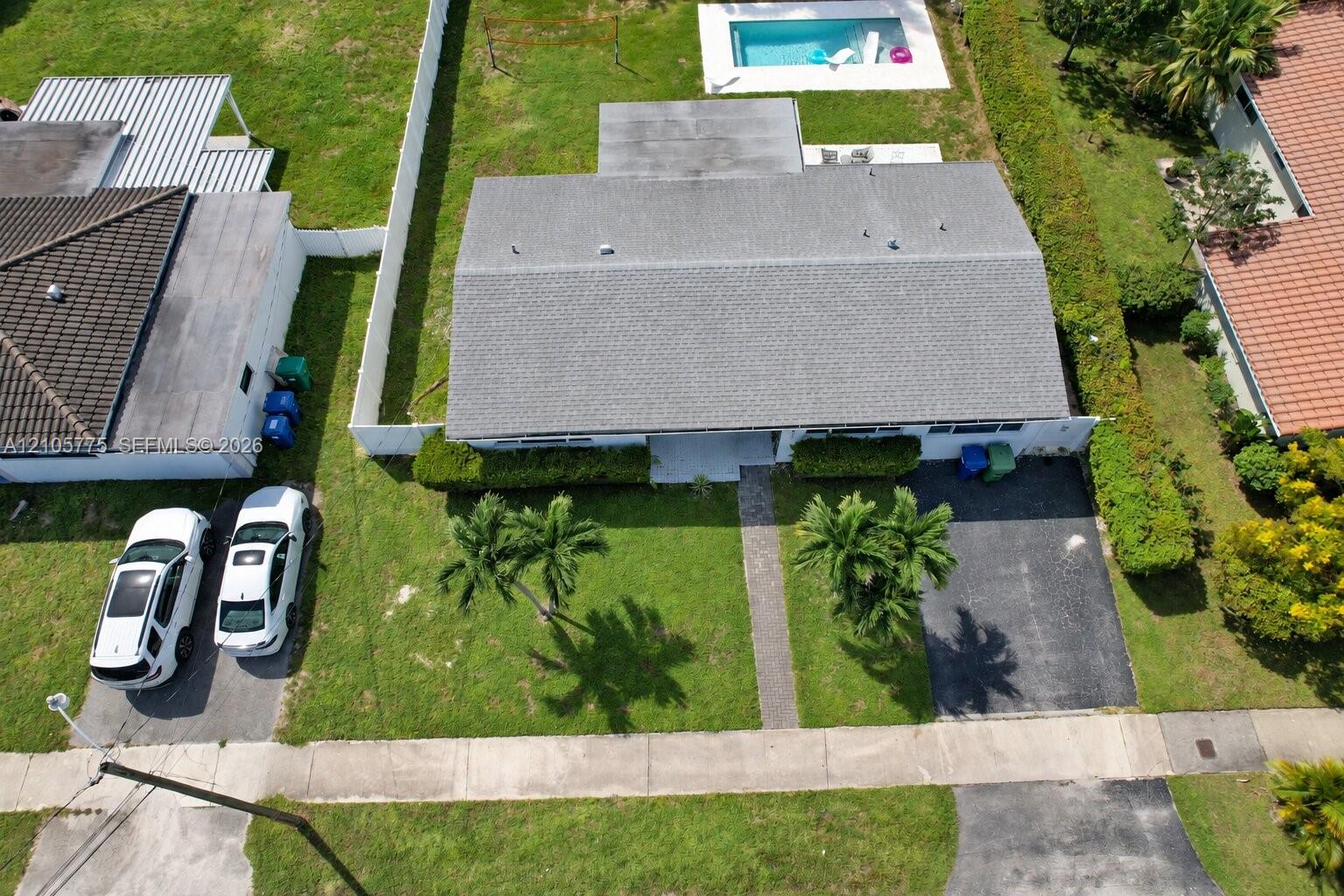 Aerial perspective featuring a shingled roof, rear swimming pool, paved driveway, concrete walkway, and landscaped lawn