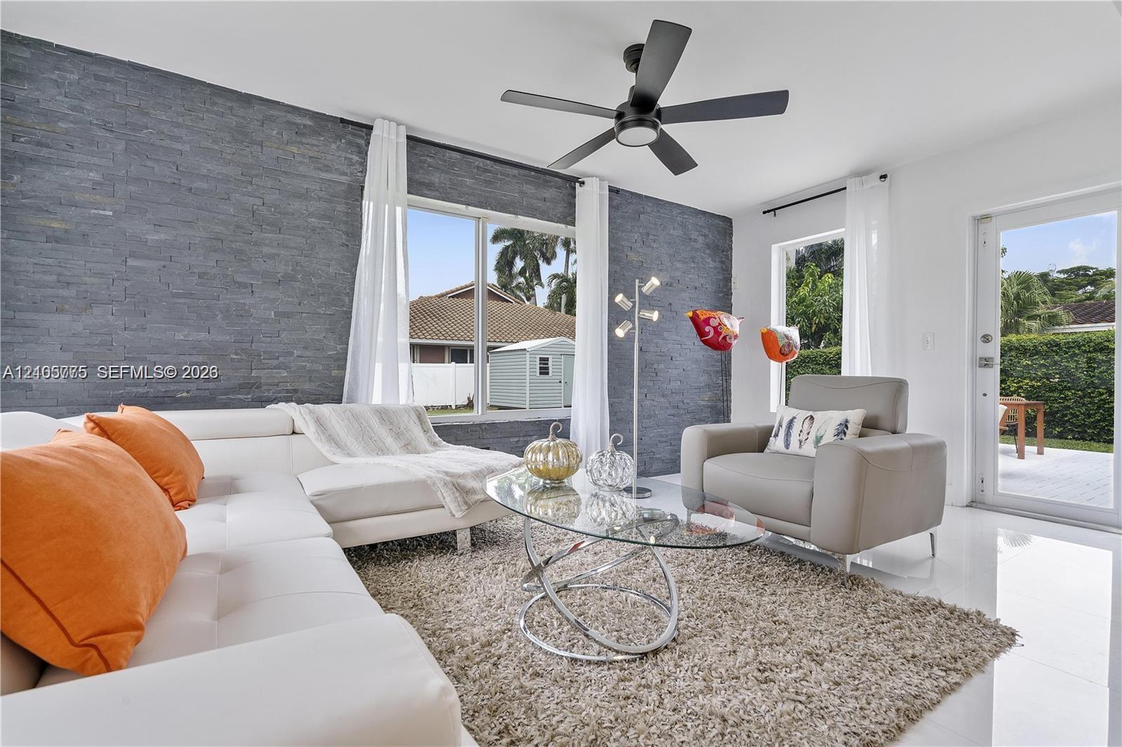 Living area featuring a floor-to-ceiling charcoal stone veneer accent wall, large picture windows, matte black ceiling fan with integrated lighting, and white ceramic tile flooring