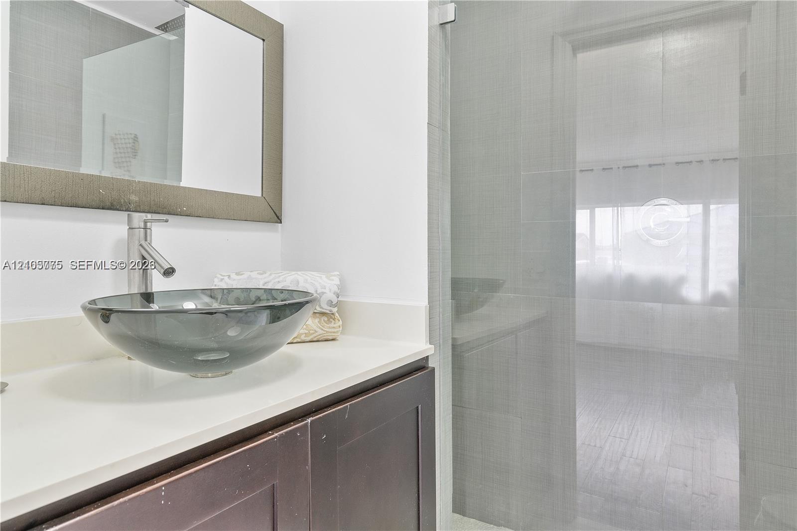 Bathroom vanity featuring a dark-finish wood cabinet, solid-surface countertop, glass vessel sink, and a framed wall mirror, accompanied by a glass-enclosed shower with tiled surround