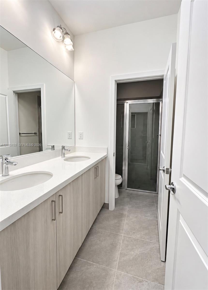 Dual-sink vanity with light-tone wood-finish cabinetry, white countertop, large wall-mounted mirror, ceramic tile flooring, and glass-enclosed shower