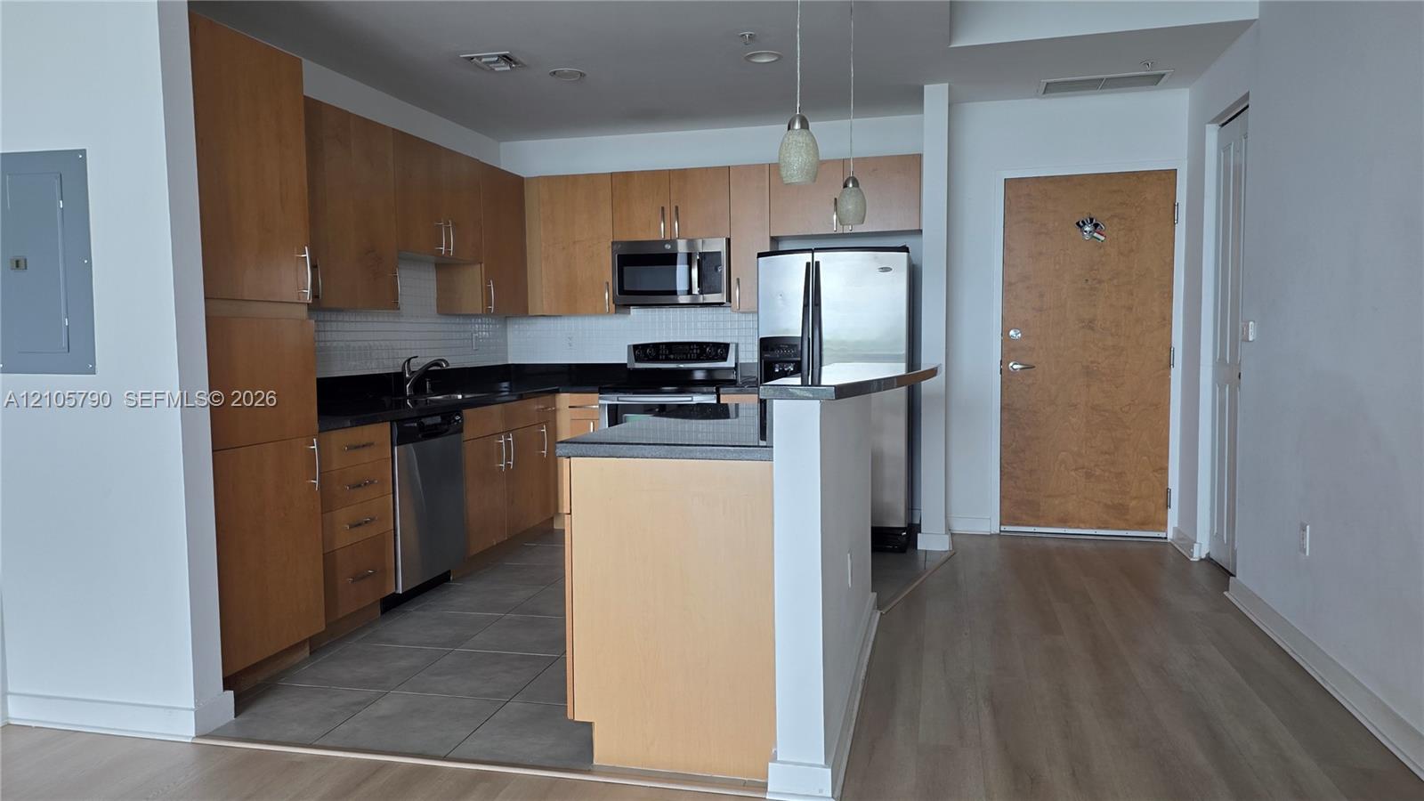 Kitchen featuring wood-finish cabinetry, dark solid-surface countertops, stainless steel appliances, tile flooring, and two pendant lights