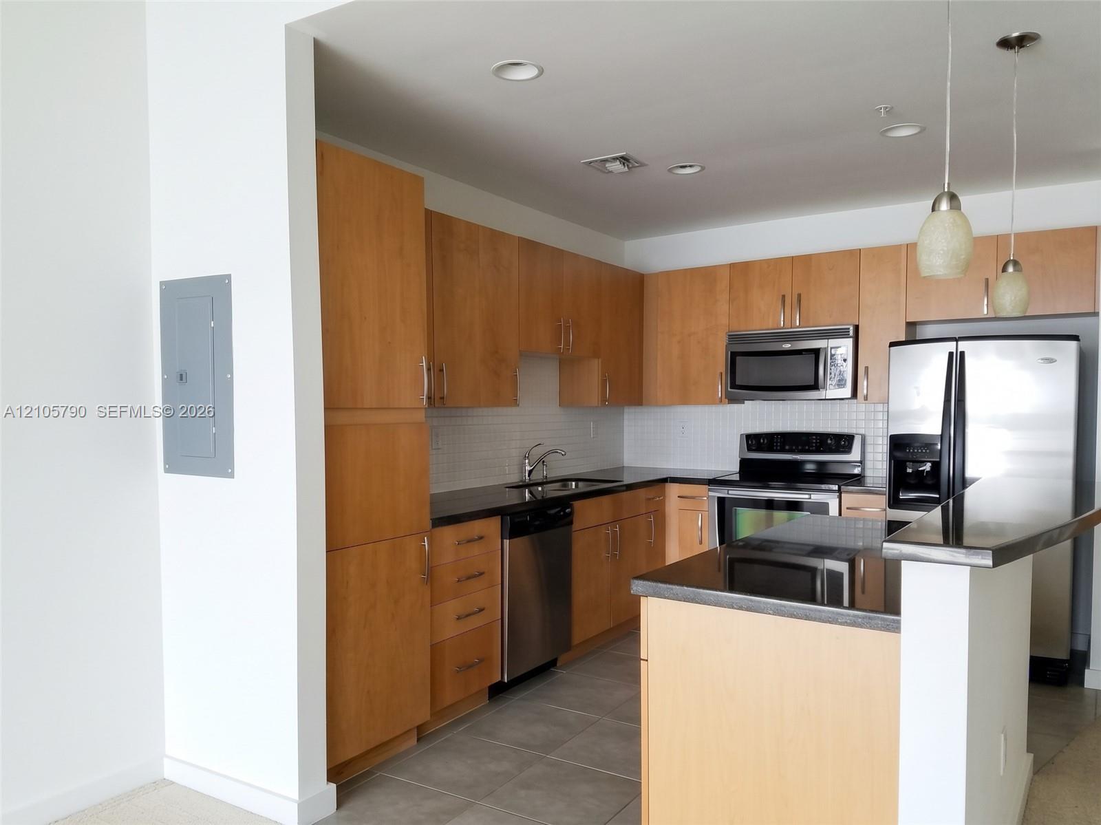 Kitchen featuring natural wood cabinetry, dark stone countertops, stainless steel appliances, tile backsplash, and two pendant lights