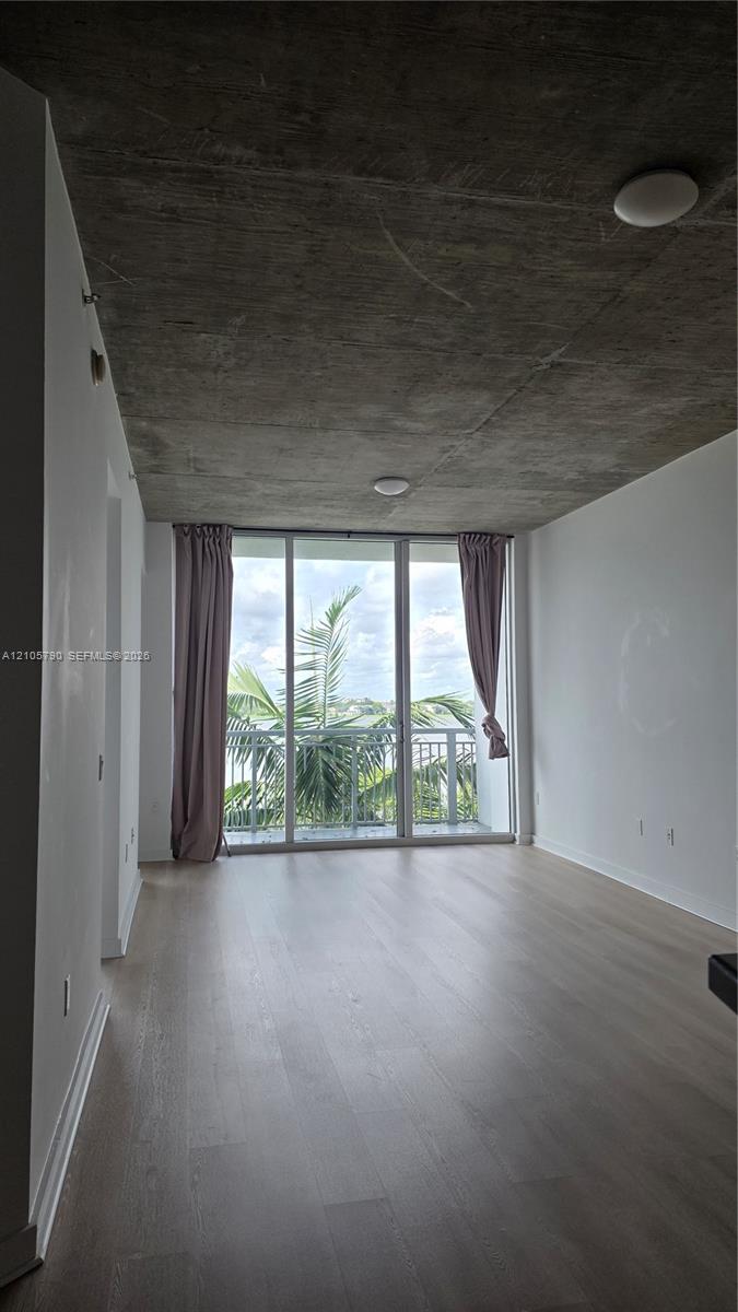 Floor-to-ceiling windows, industrial-style exposed concrete ceiling, light-toned wood-finish flooring, and balcony access