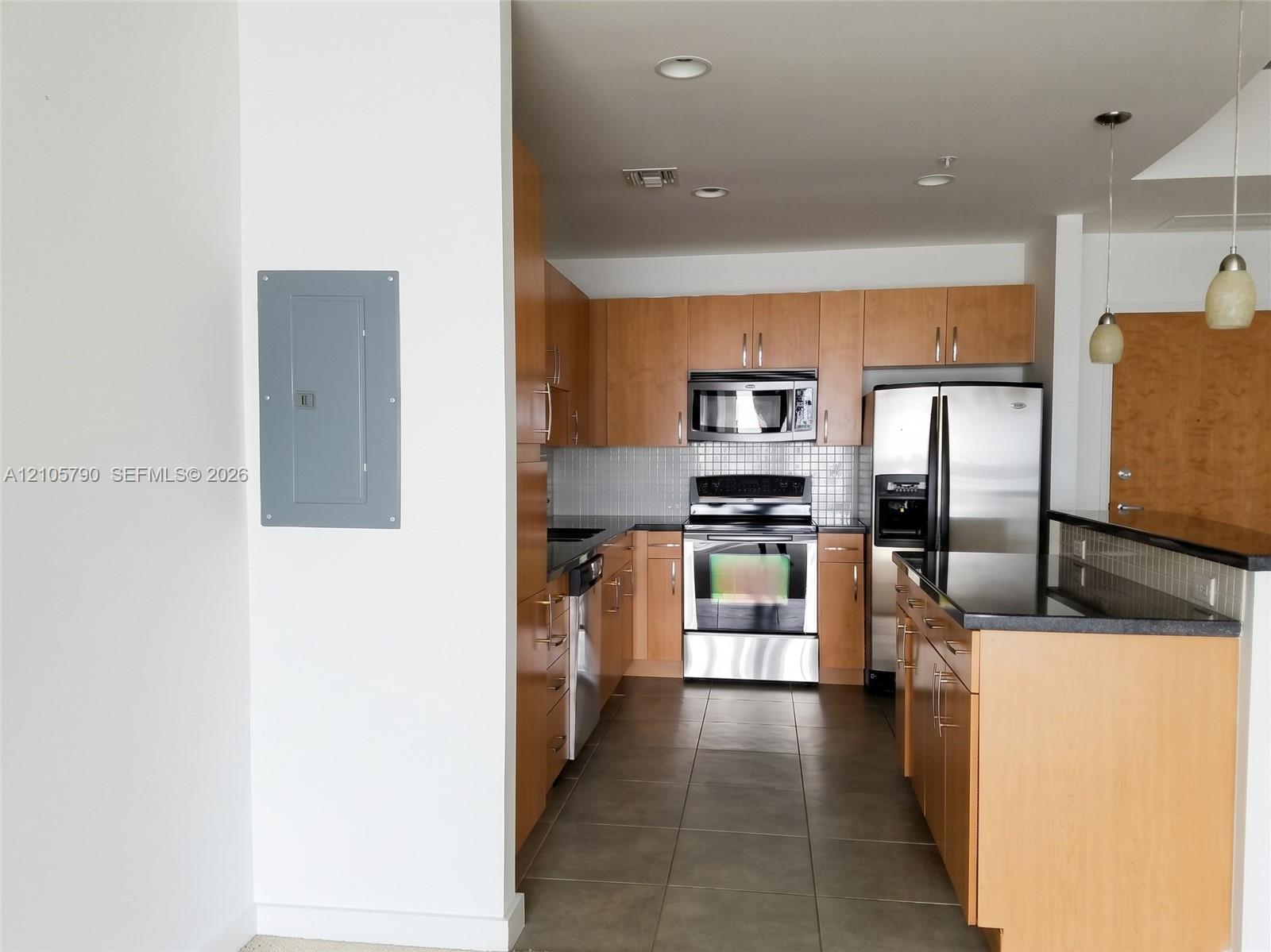 Kitchen featuring light wood-finish cabinetry, stainless steel appliances, dark stone countertops, tile backsplash, and oversized ceramic floor tiles