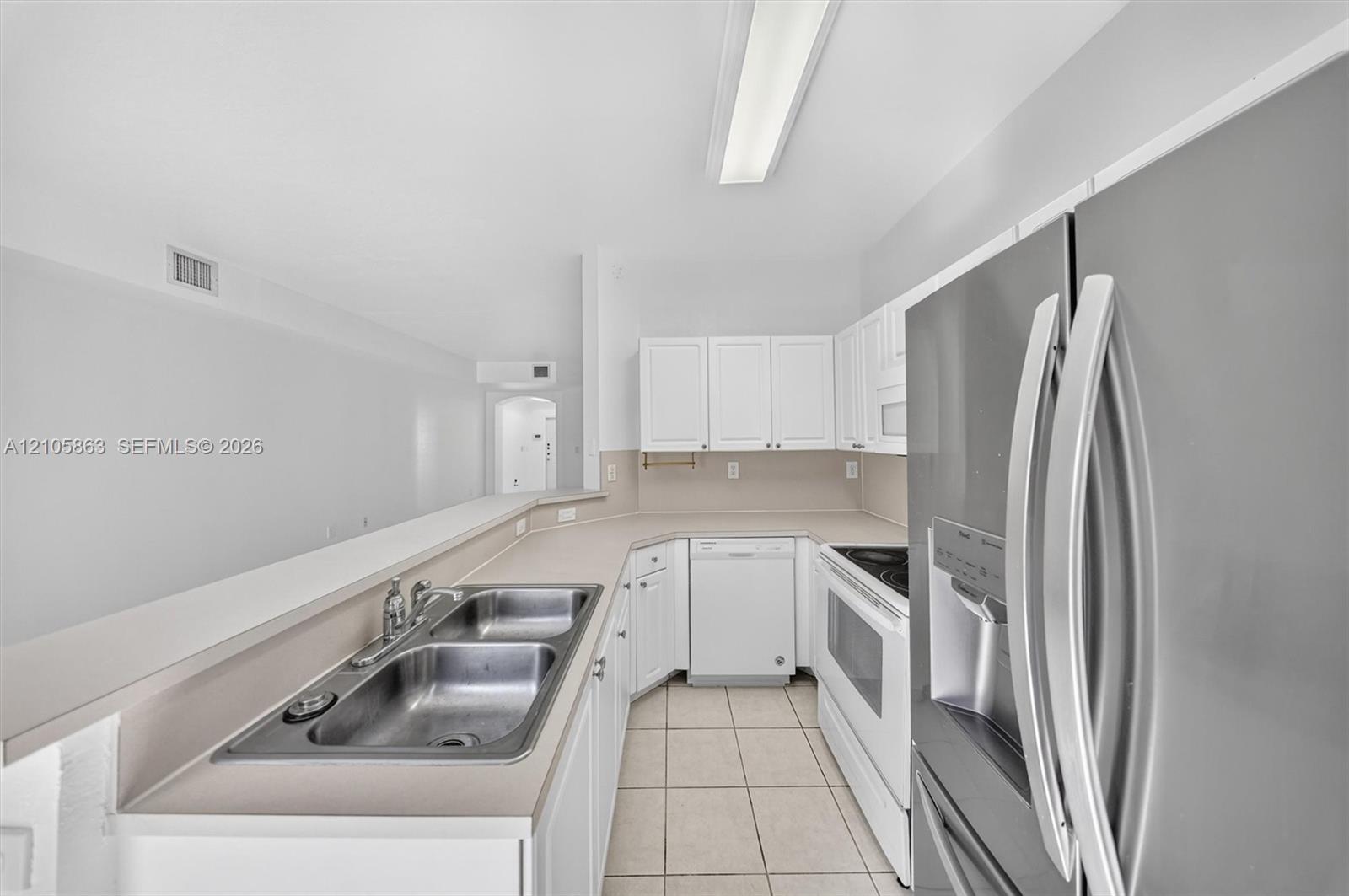Kitchen featuring white cabinetry, solid-surface countertops, dual-basin stainless steel sink, tile flooring, and stainless steel refrigerator