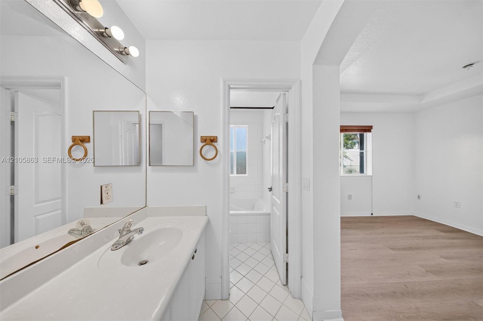 Full-width vanity with integrated sink, expansive wall-to-wall mirror, brushed metal fixtures, white tiled flooring, and wood-finish flooring