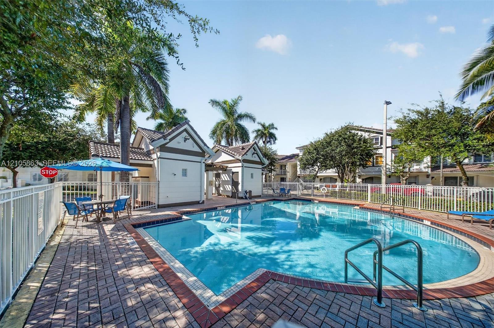 In-ground swimming pool with brick paver decking, white perimeter fencing, and two pool-side structures featuring pitched rooflines