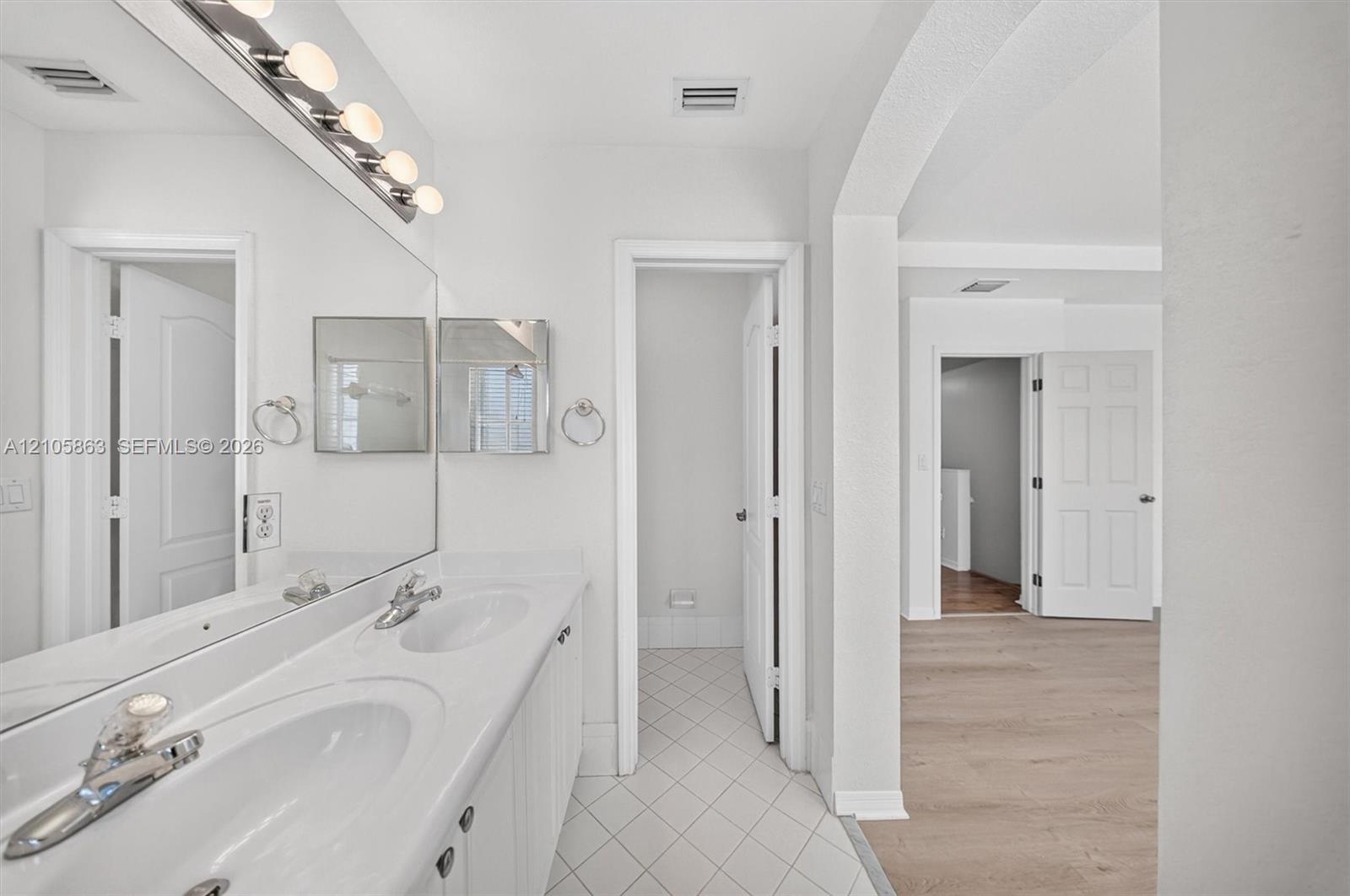 Dual-sink vanity with white countertop, full-width vanity mirror, polished chrome fixtures, white tile flooring, and arched architectural detailing