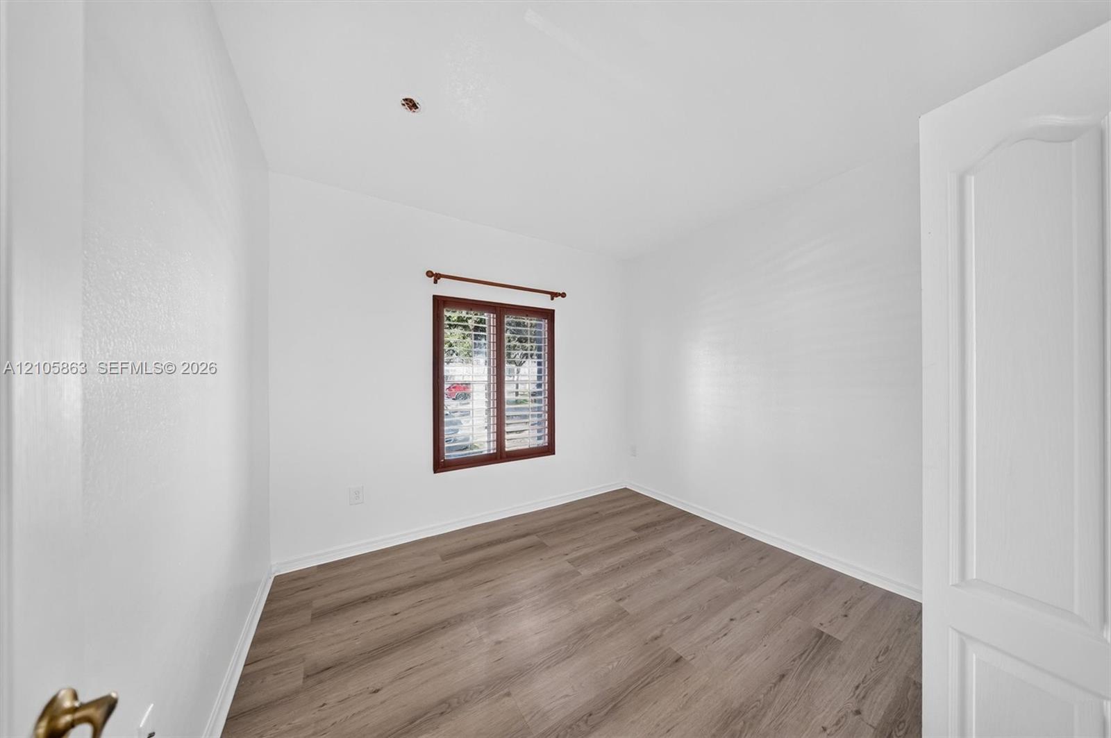 Room interior featuring wood-finish flooring, white walls, and a window equipped with plantation shutters and a wooden curtain rod