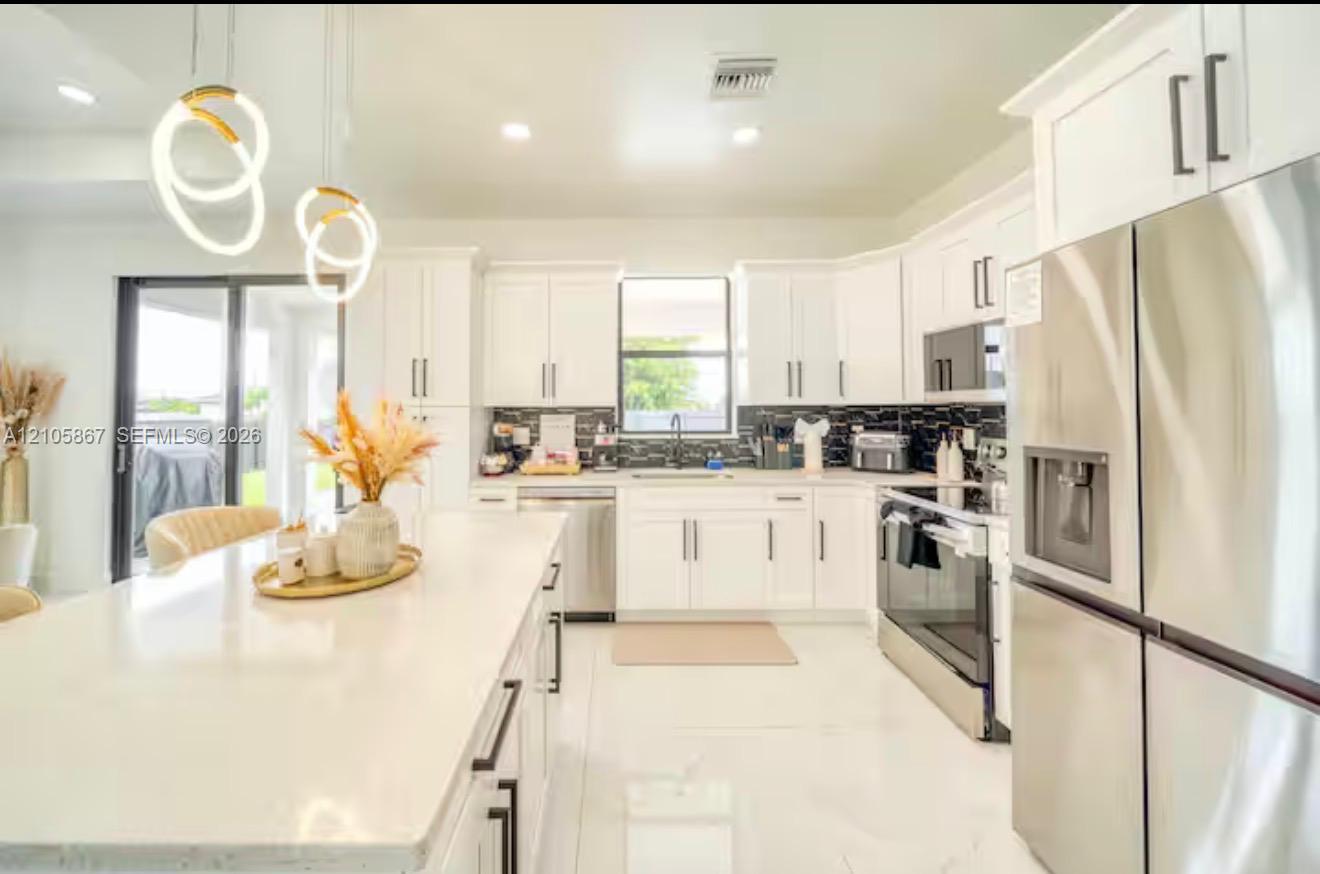Kitchen featuring white shaker-style cabinetry, a quartz waterfall island, stainless steel appliances, a dark tile backsplash, and contemporary sculptural pendant lighting