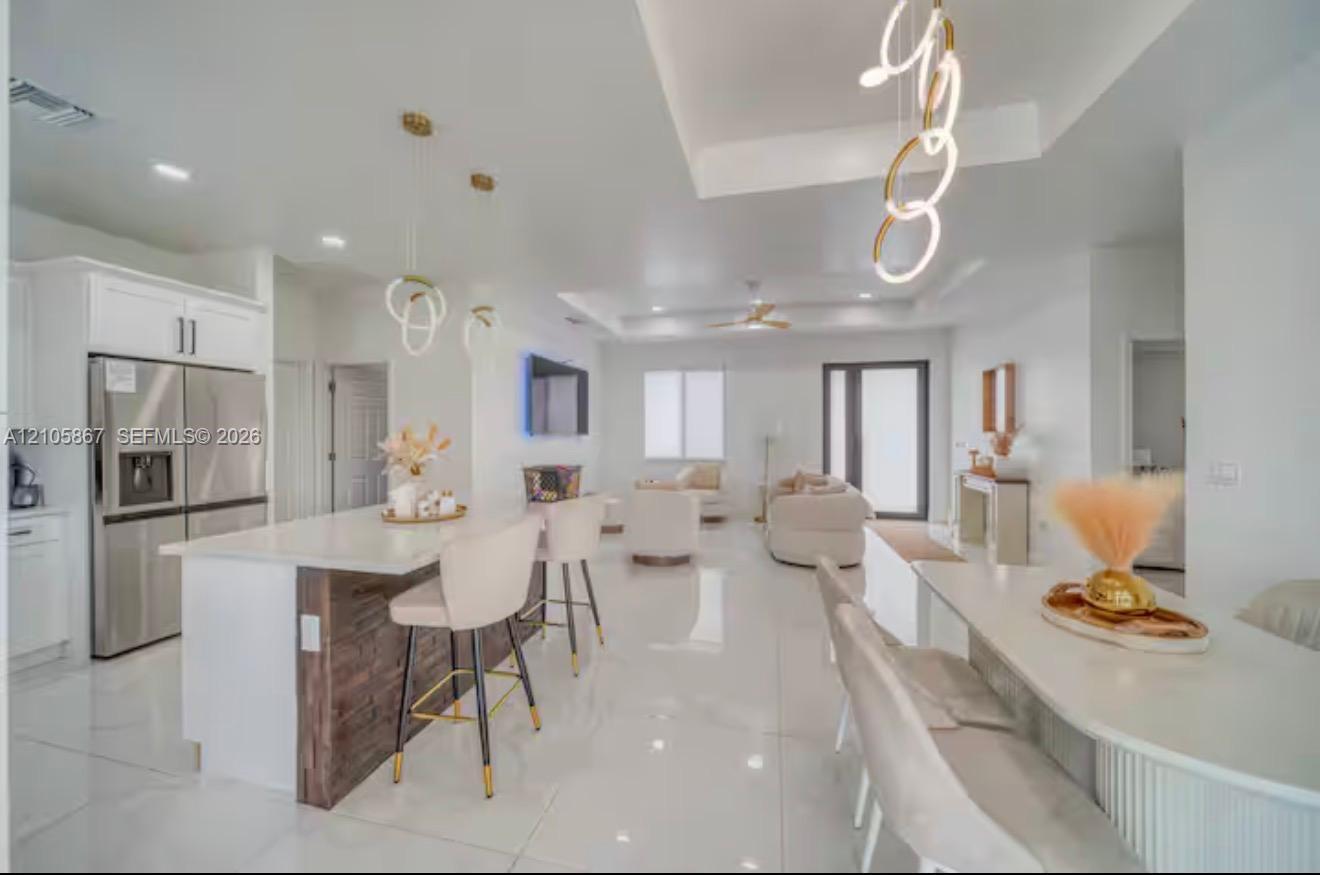 Open-concept living space featuring high-gloss tile flooring, white shaker-style cabinetry, a kitchen island with stone-finish countertops and wood-accented base, tray ceilings, and contemporary ring-style pendant lighting