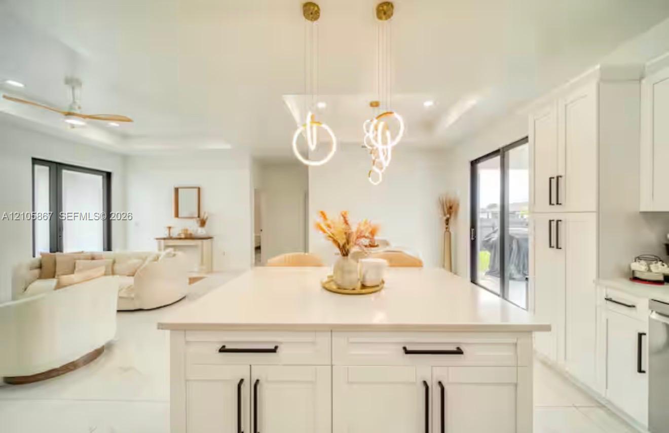 Kitchen featuring an expansive center island with solid-surface countertop, white Shaker-style cabinetry, matte black hardware, and contemporary ring pendant lighting Kitchen featuring an expansive center island with solid-surface countertop, white Shaker-style cabinetry, matte black hardware, and contemporary ring pendant lighting