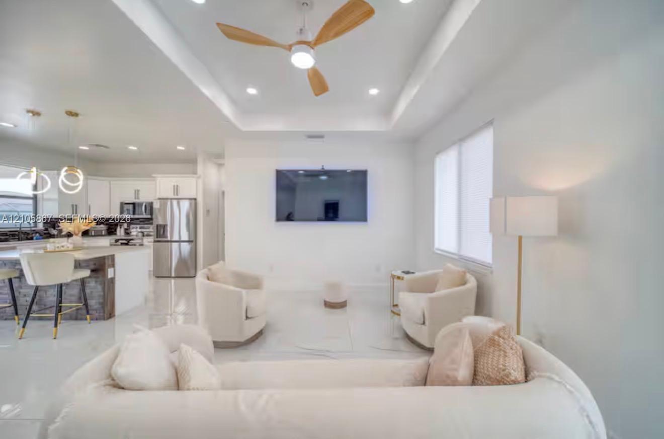 Open-concept interior featuring a recessed tray ceiling, three-blade wood-finish ceiling fan, recessed lighting, white tile flooring, and a kitchen island with stone-finish base Open-concept interior featuring a recessed tray ceiling, three-blade wood-finish ceiling fan, recessed lighting, white tile flooring, and a kitchen island with stone-finish base