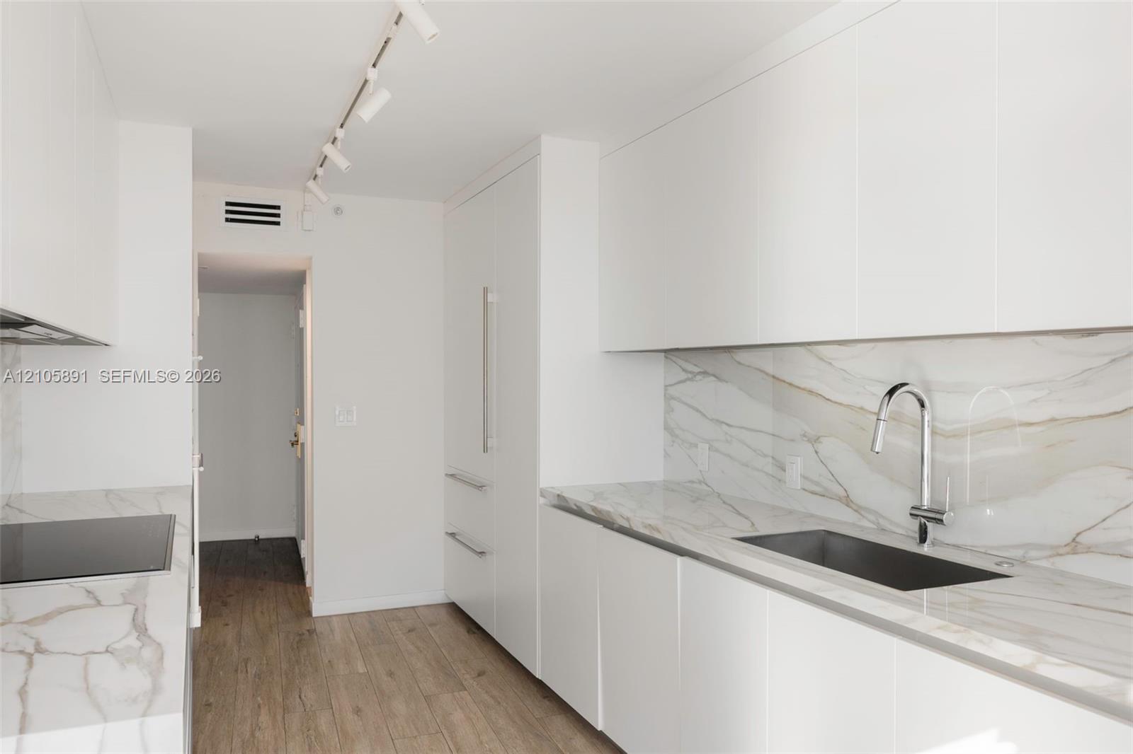 Modern kitchen featuring white flat-panel cabinetry, stone-finish countertops and backsplash, integrated refrigerator, undermount sink with high-arc faucet, and wood-finish flooring