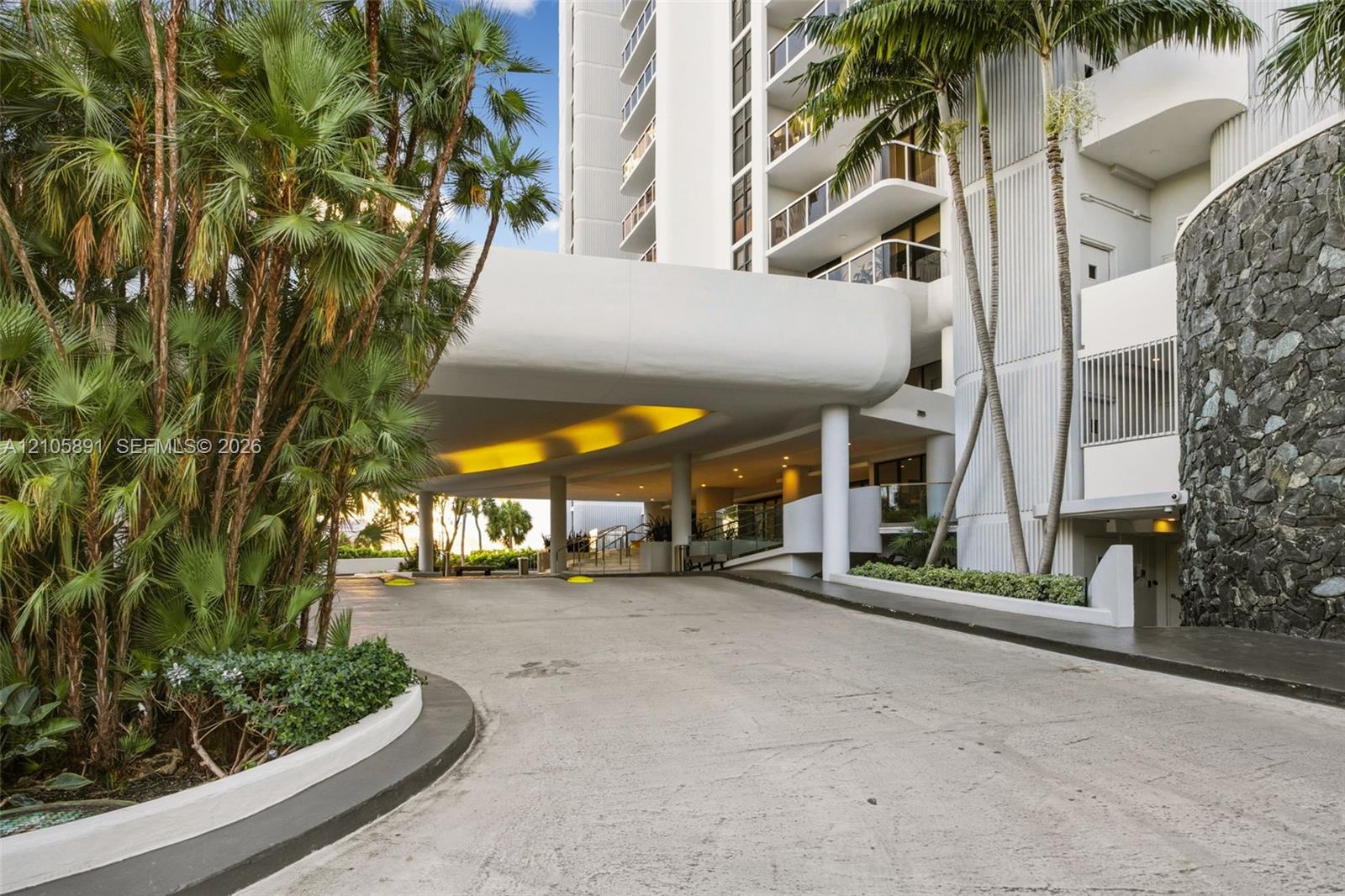 Covered porte-cochre entry featuring stone masonry accents, a concrete drive, and mature palm landscaping