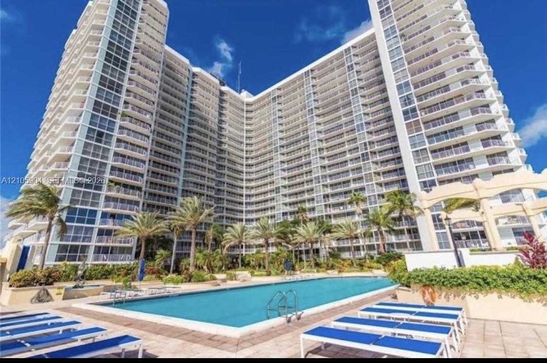 High-rise building exterior featuring a multi-story facade with wraparound balconies, accompanied by an outdoor swimming pool, paved sun deck, and mature palm trees High-rise building exterior featuring a multi-story facade with wraparound balconies, accompanied by an outdoor swimming pool, paved sun deck, and mature palm trees