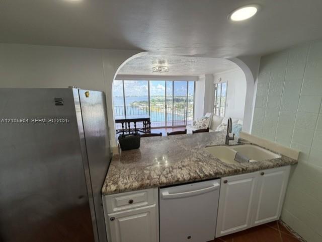 Kitchen featuring stone countertops, white cabinetry, stainless steel refrigerator, white dishwasher, and double-basin undermount sink with arched opening