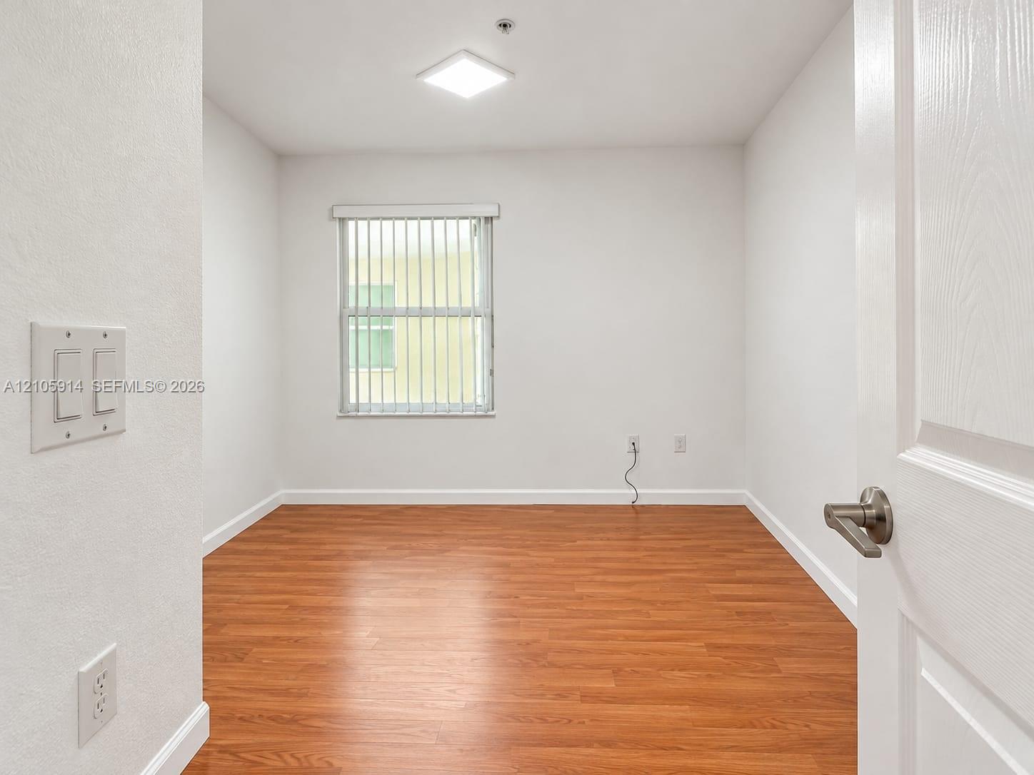 Room featuring wood-finish flooring, a flush-mount light fixture, and a window with vertical blinds