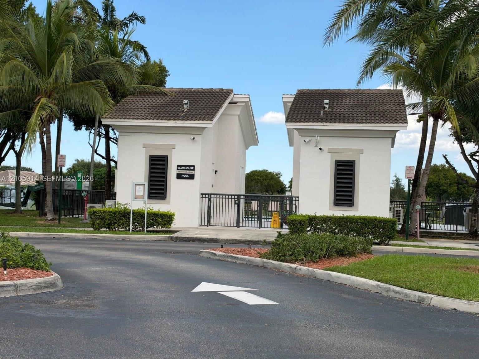 Gated entry featuring dual stucco structures, tile roofing, and metal security fencing surrounded by manicured landscaping and palm trees