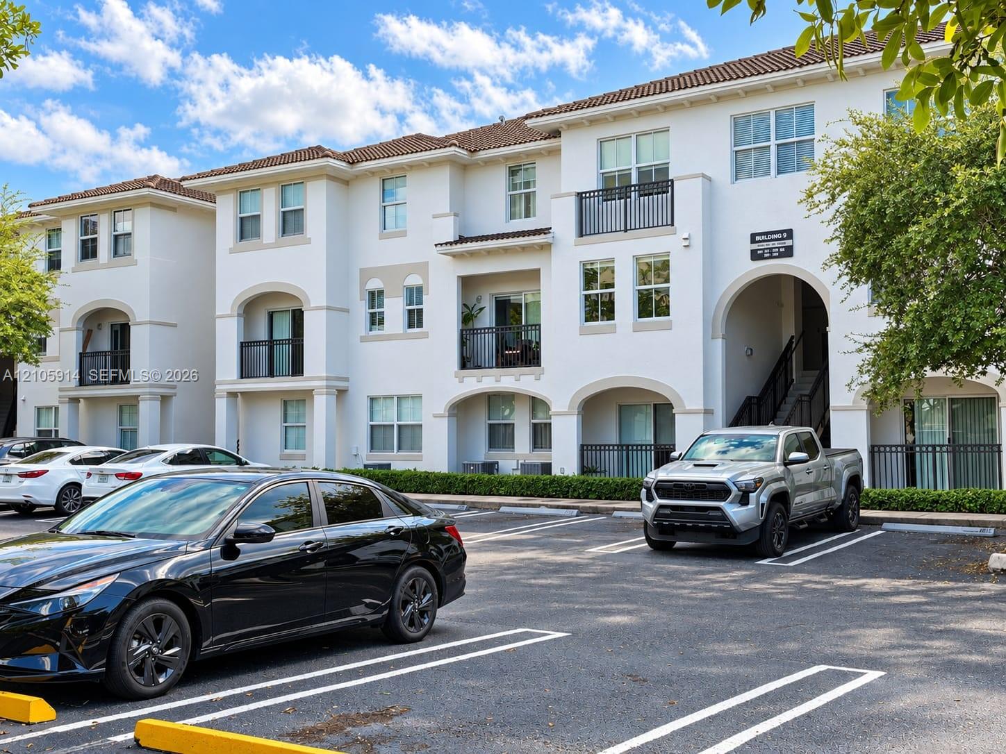 Multi-story stucco exterior featuring arched architectural detailing, tile roof, private balconies with metal railings, and surface parking