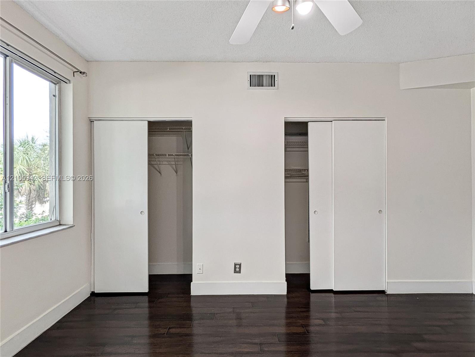 Dark wood-finish flooring, dual closets with sliding panel doors, ceiling fan with integrated lighting, and a window with exterior greenery